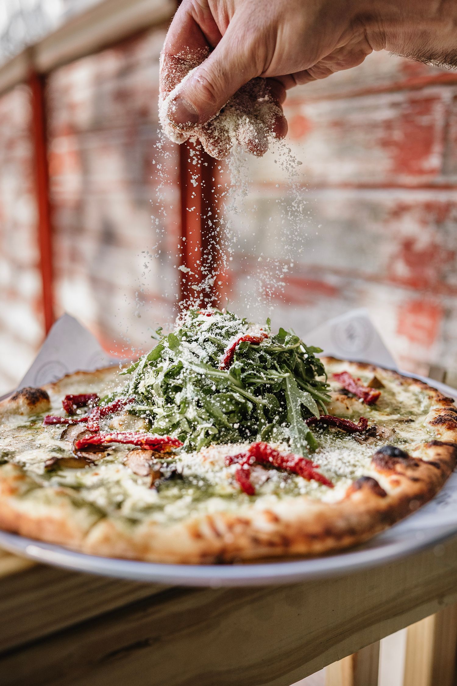 A hand sprinkling parmesan cheese over a fresh pizza topped with arugula and sun-dried tomatoes on a wooden table.