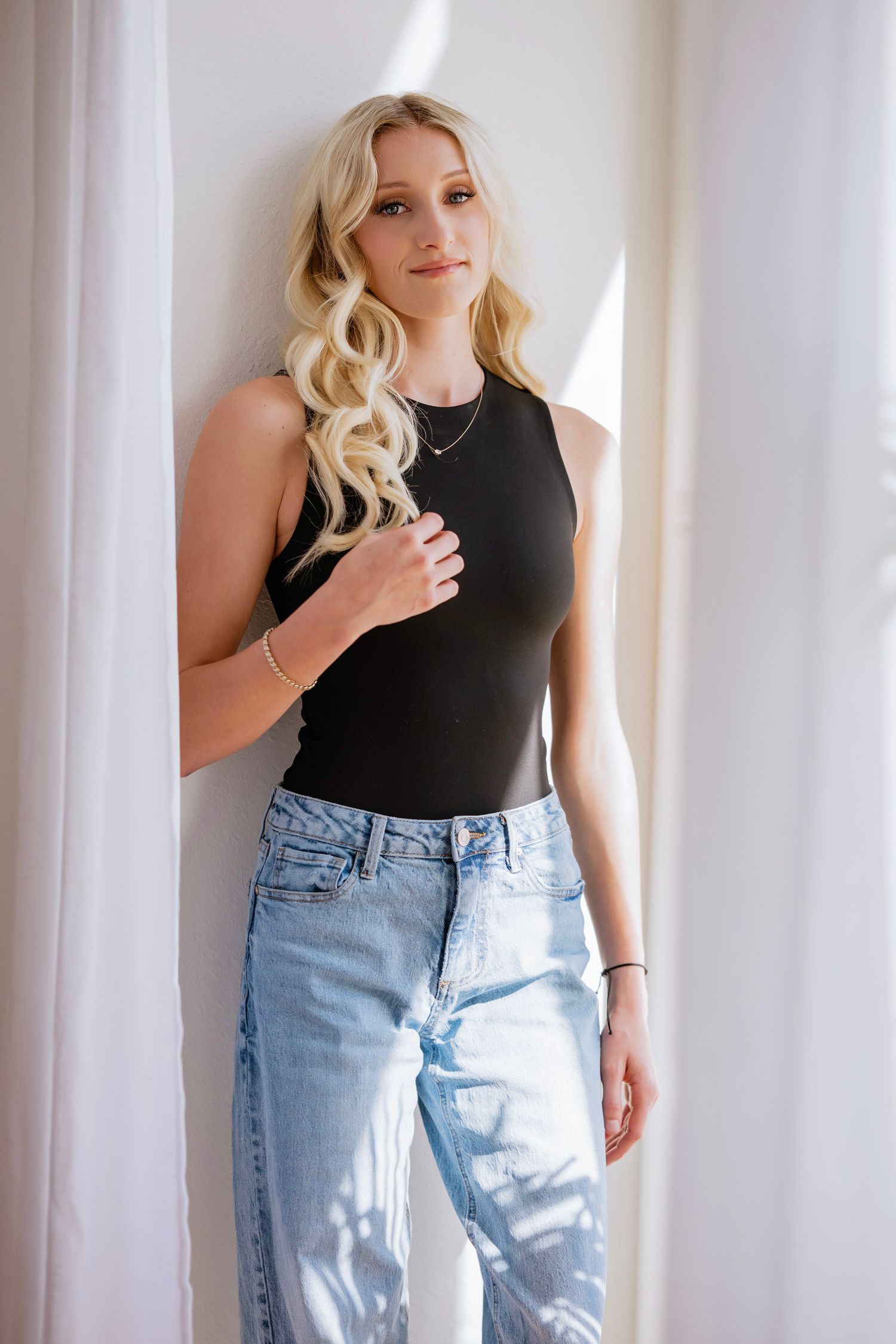 Blonde woman in black sleeveless bodysuit and light blue jeans leaning against a white wall.