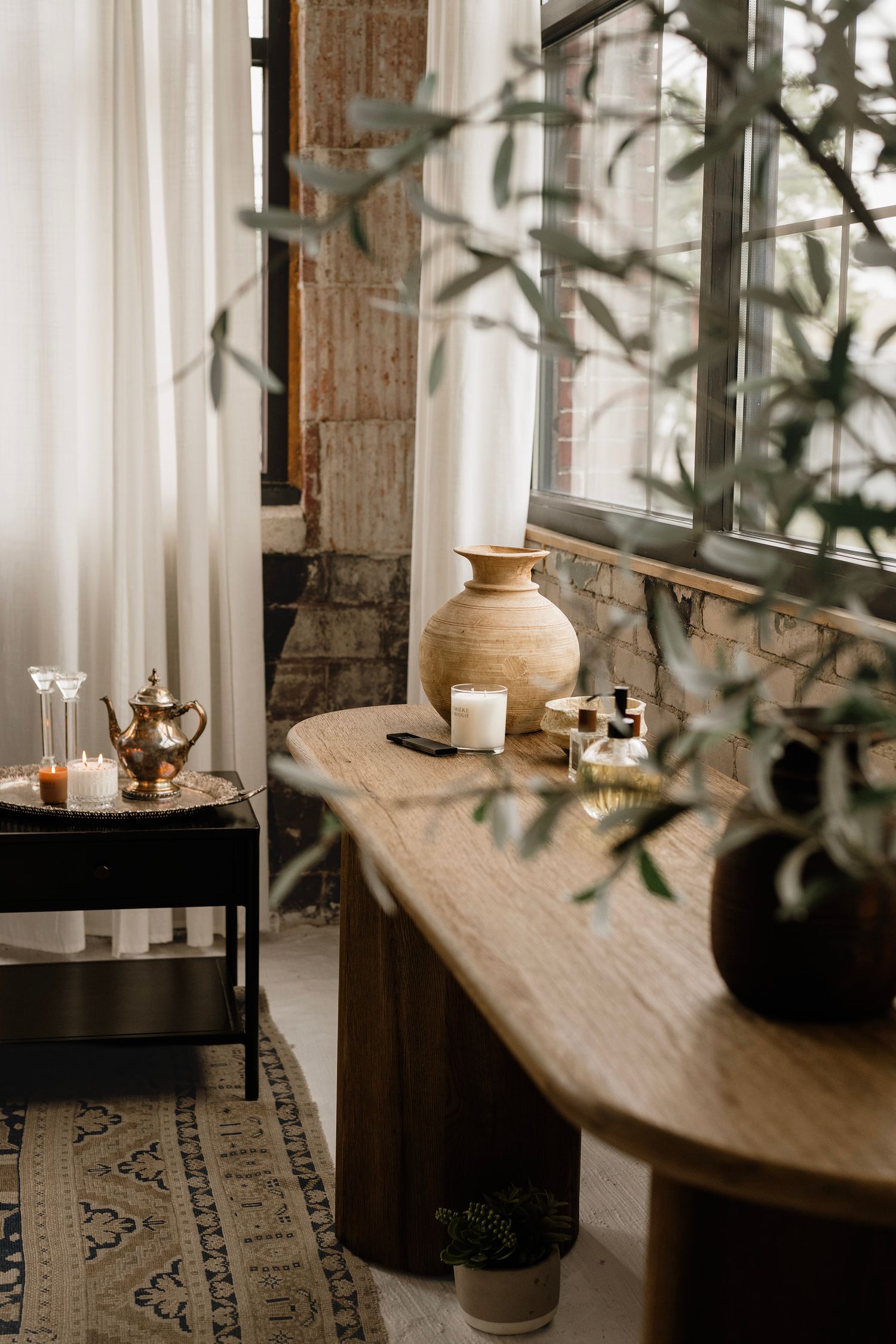 Rustic wooden bench with ceramic vase, candles, and botanicals near window with sheer curtains in boho interior.
