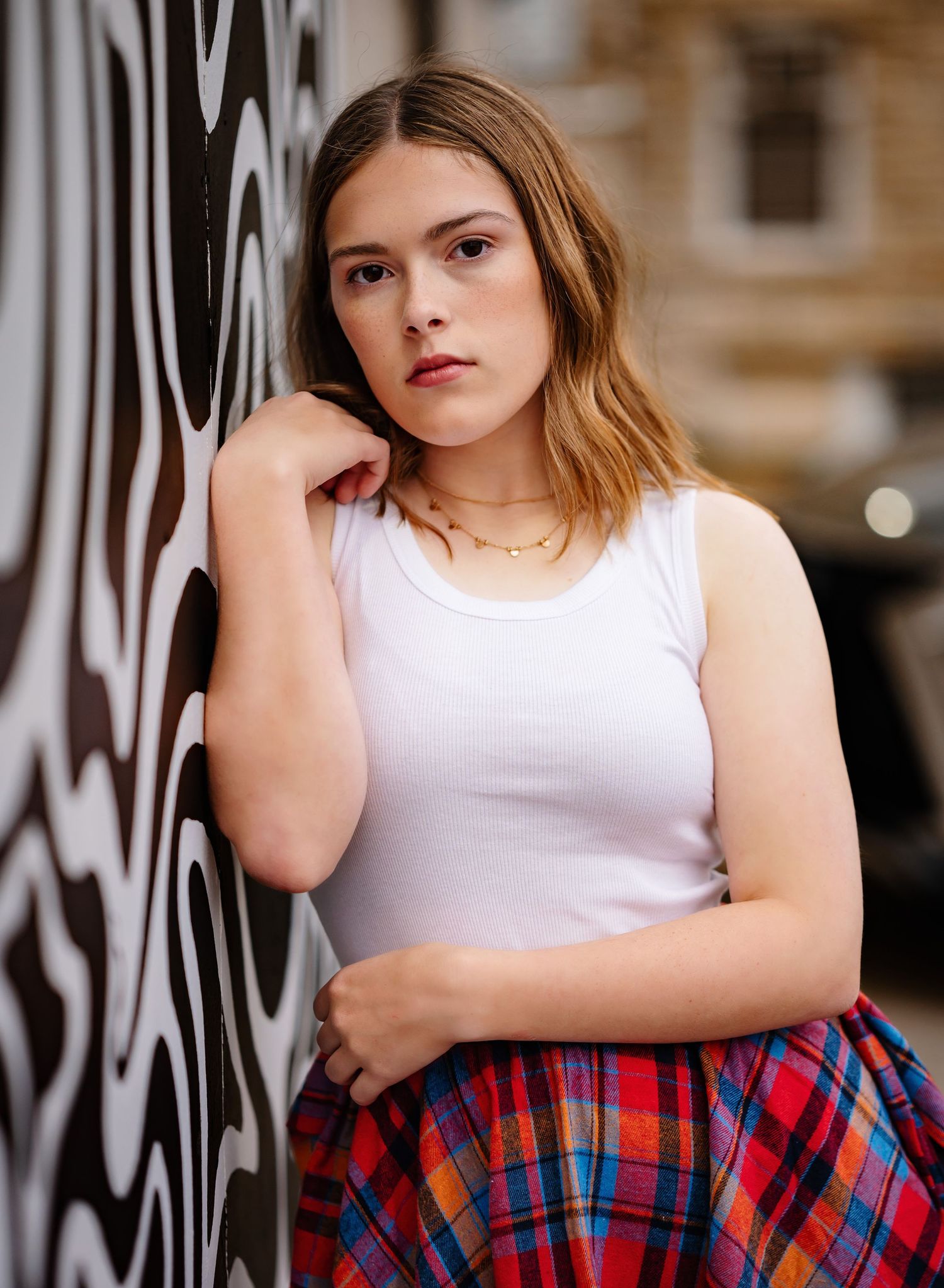 Person in white tank top and plaid shirt leaning against wall with graffiti in urban setting.