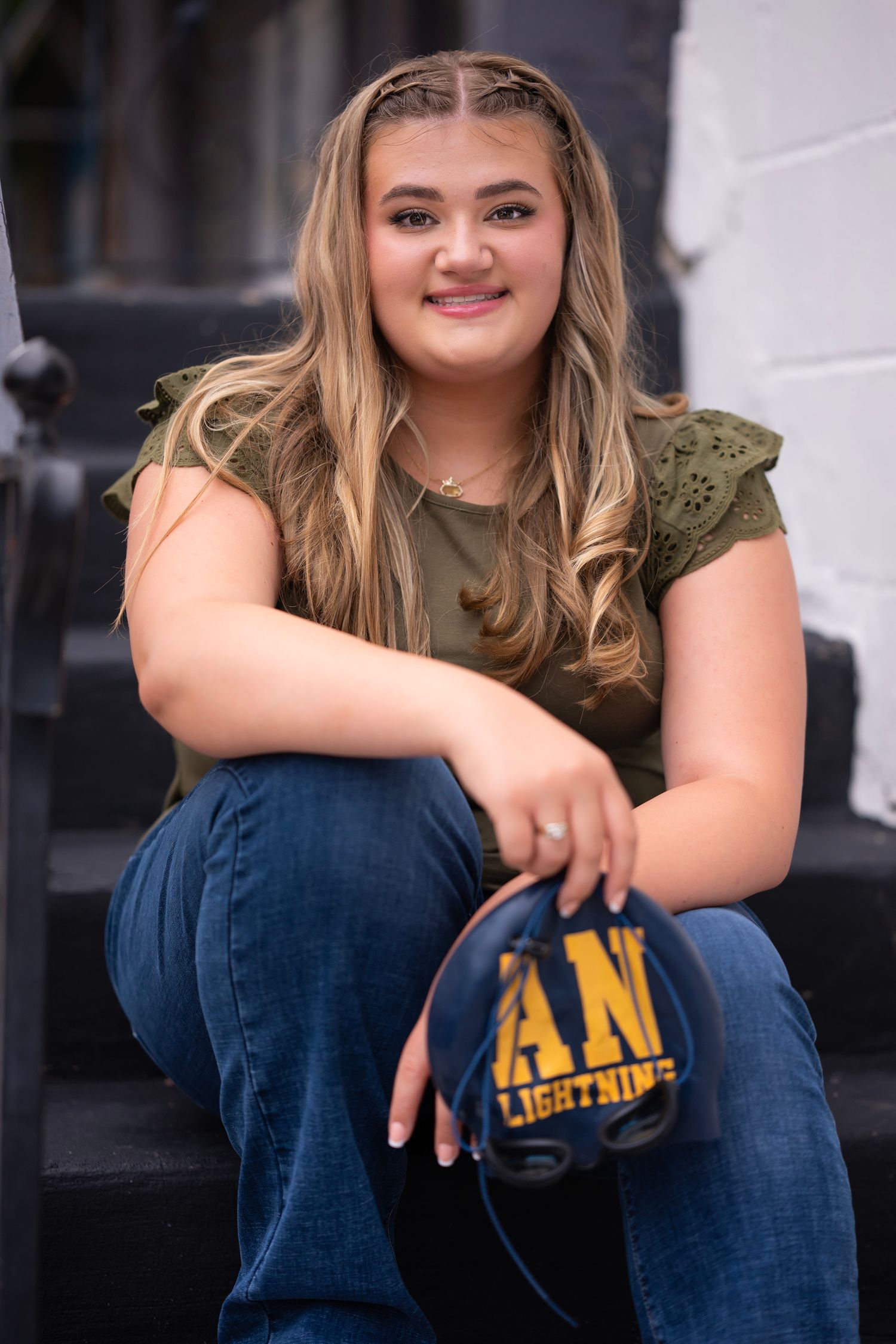 Young woman sitting on steps smiling, wearing olive green top and jeans, holding a Lightning cap.