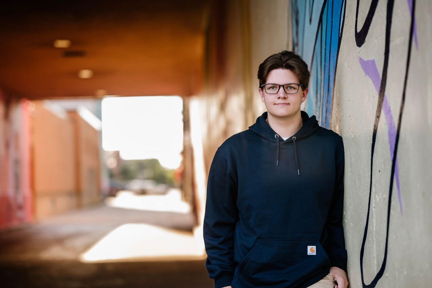 A person in glasses and dark clothing stands against a wall with blue accents in an indoor setting.
