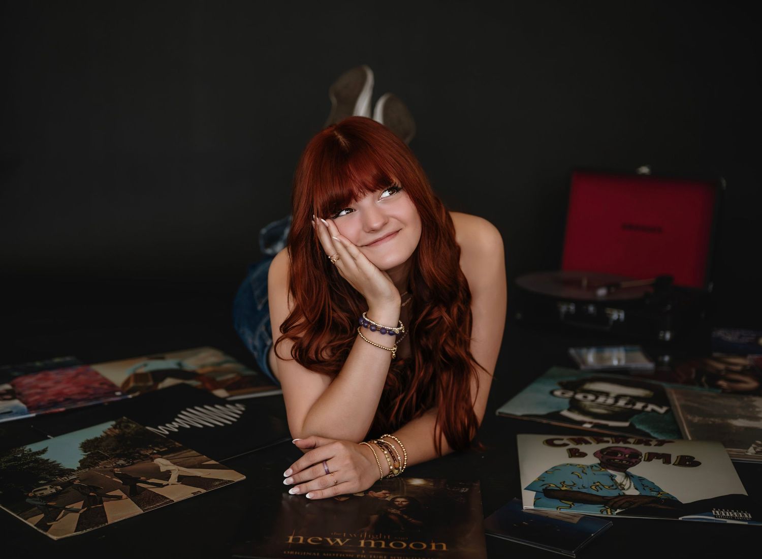 A person with long red hair lies on their stomach surrounded by vinyl records in a dark room.