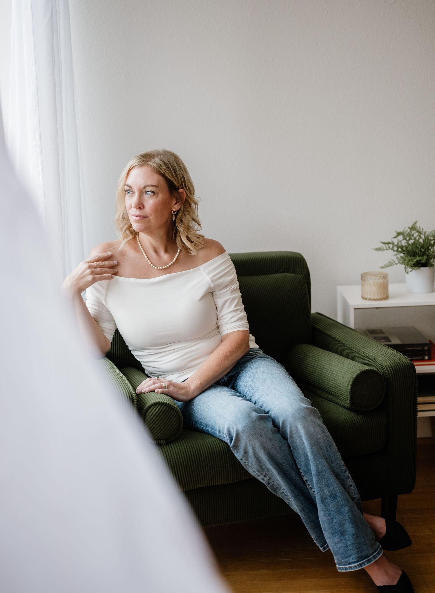 A relaxed person in jeans and white top lounges casually on a dark green sofa near a bright window.