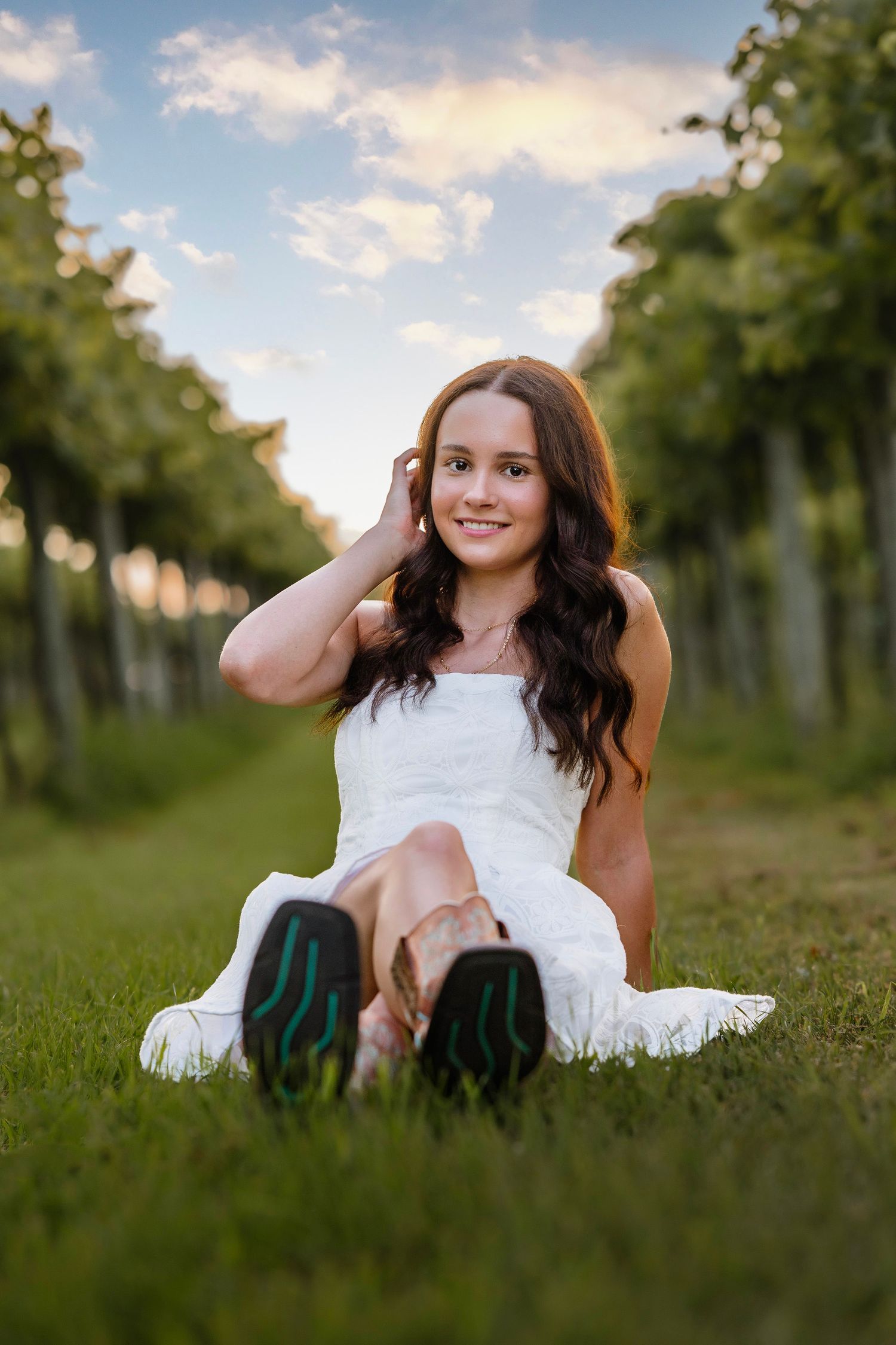 Young woman in white dress sitting on grass between vineyard rows at sunset, smiling with hand in hair.