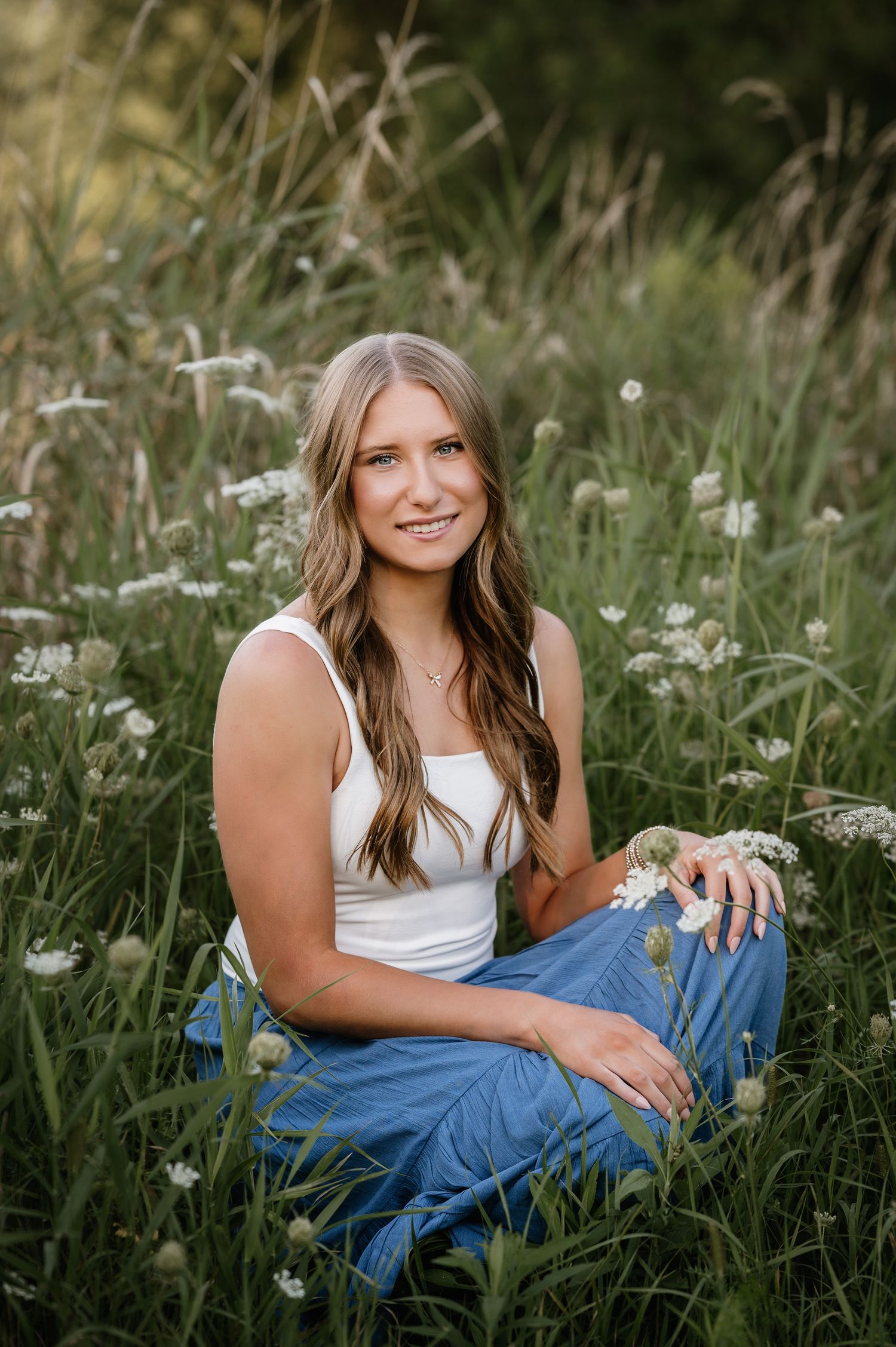 Young woman sitting in a wildflower meadow wearing a white tank top and blue wide-leg pants, smiling softly.