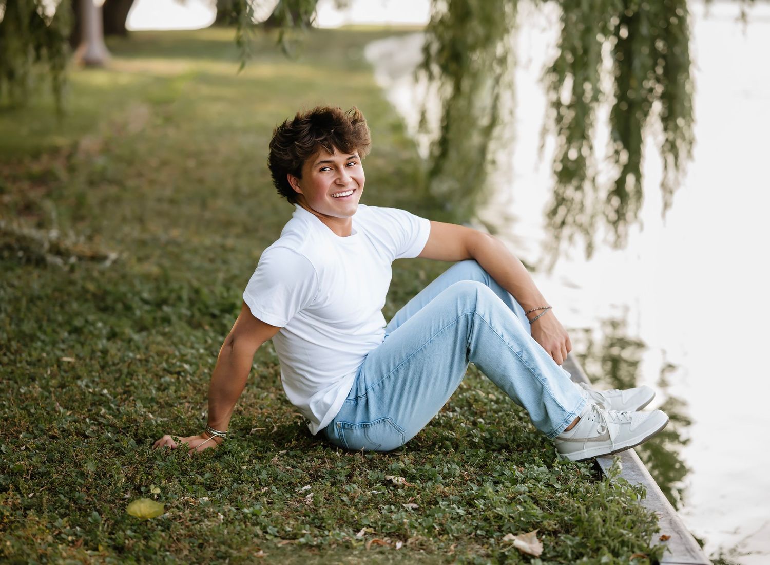 Someone in a white t-shirt and jeans relaxes on grassy ground beneath willow trees on a sunny day.