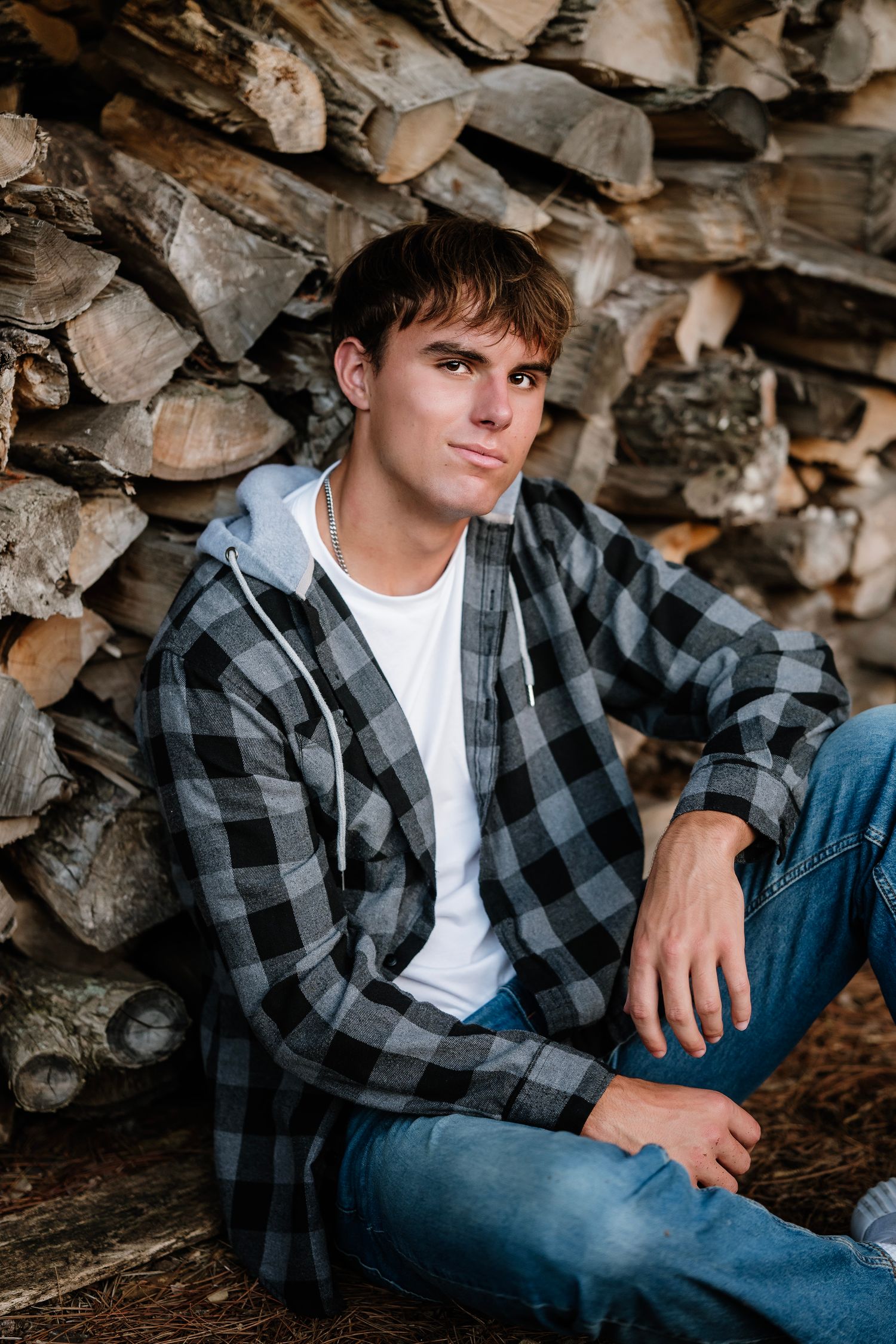 A person in a black and gray plaid flannel shirt and jeans poses against a woodpile background.