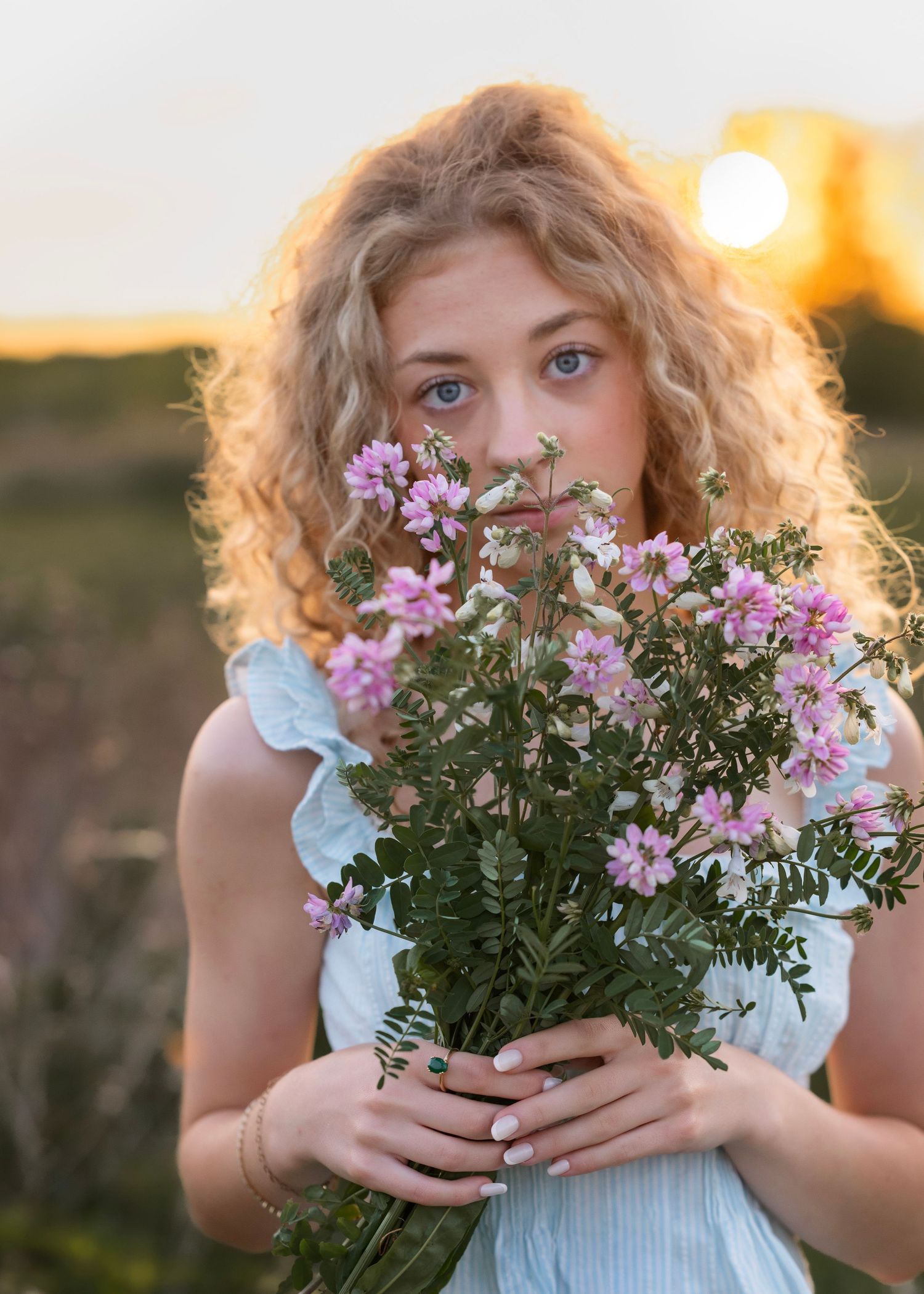 Young woman with curly blonde hair holding pink wildflowers at golden hour sunset in a field.
