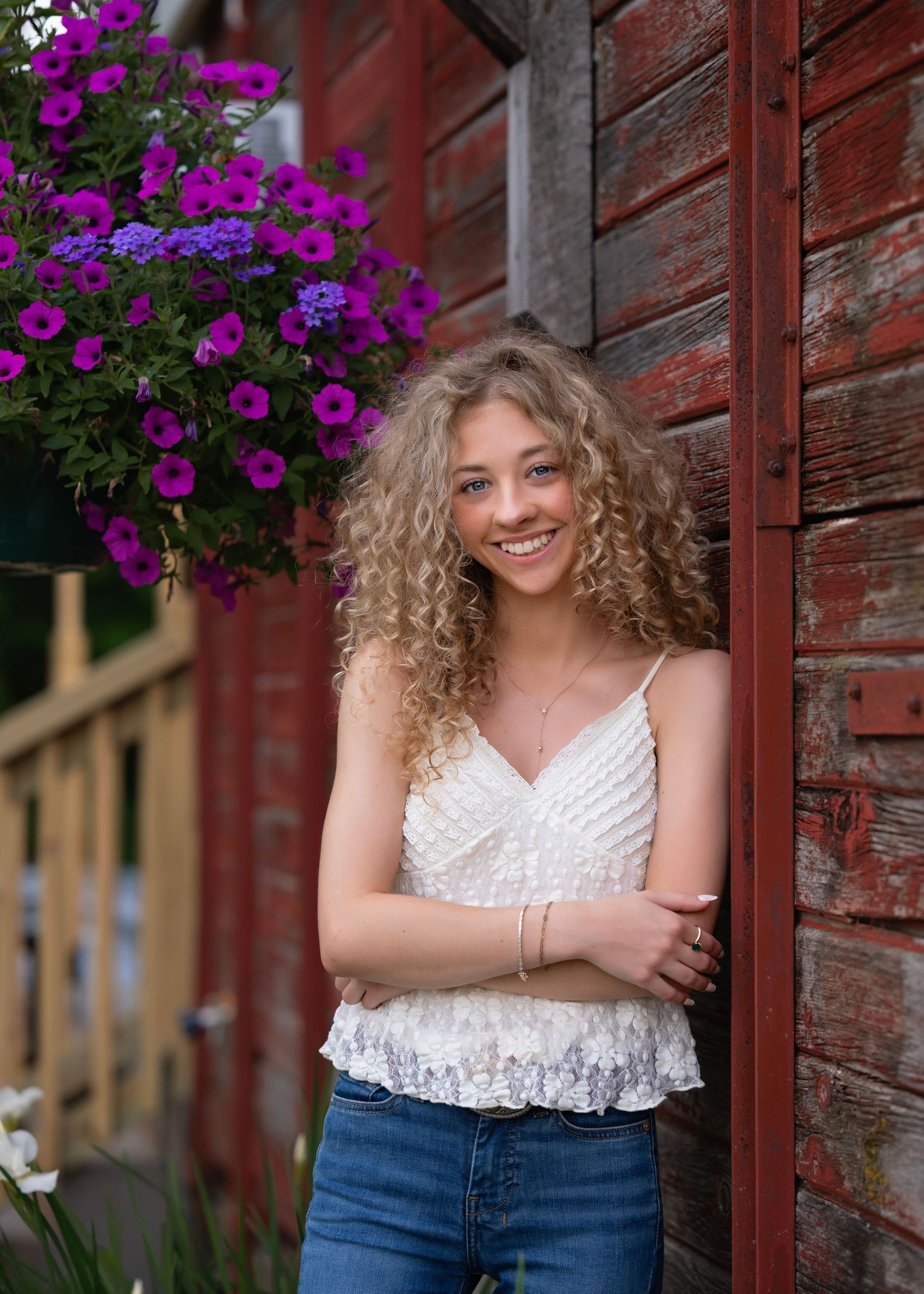 Smiling teenage girl with curly blonde hair in white lace top and jeans leaning against red barn wall with purple flowers.