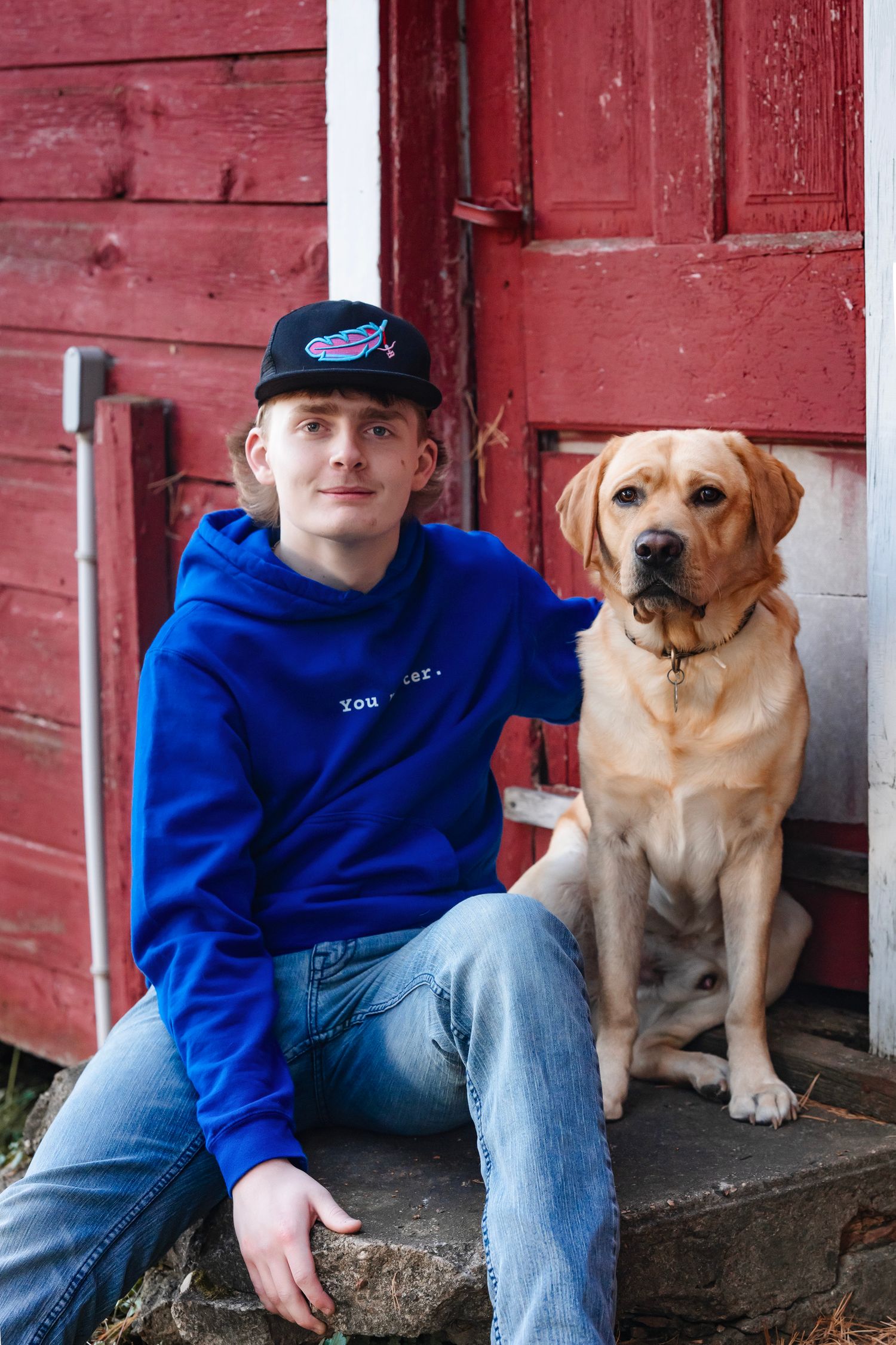 A yellow Labrador retriever sits next to someone wearing a blue hoodie and baseball cap on the steps of a red barn.