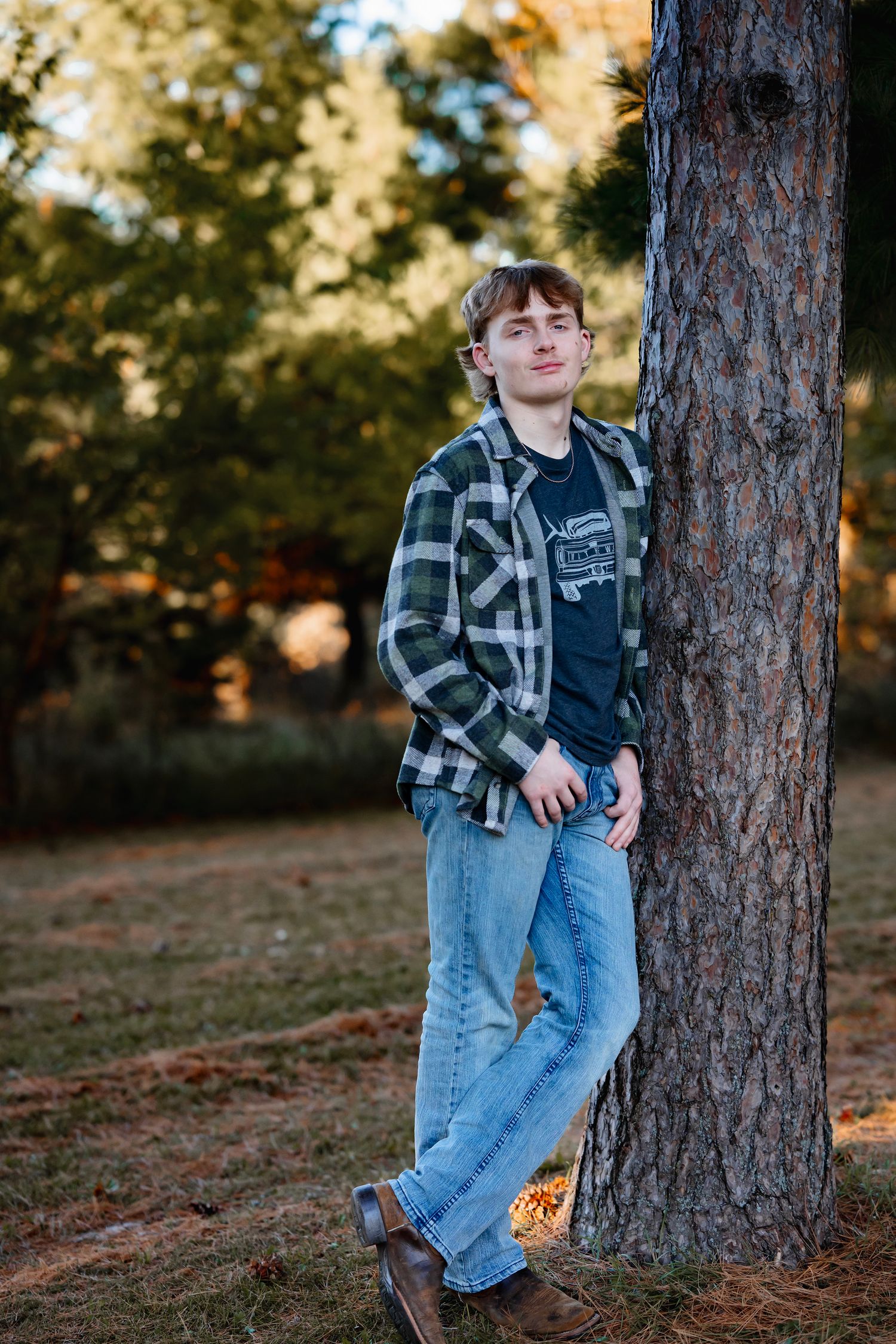 Child in plaid flannel and jeans poses casually against a tree trunk during golden hour in autumn.