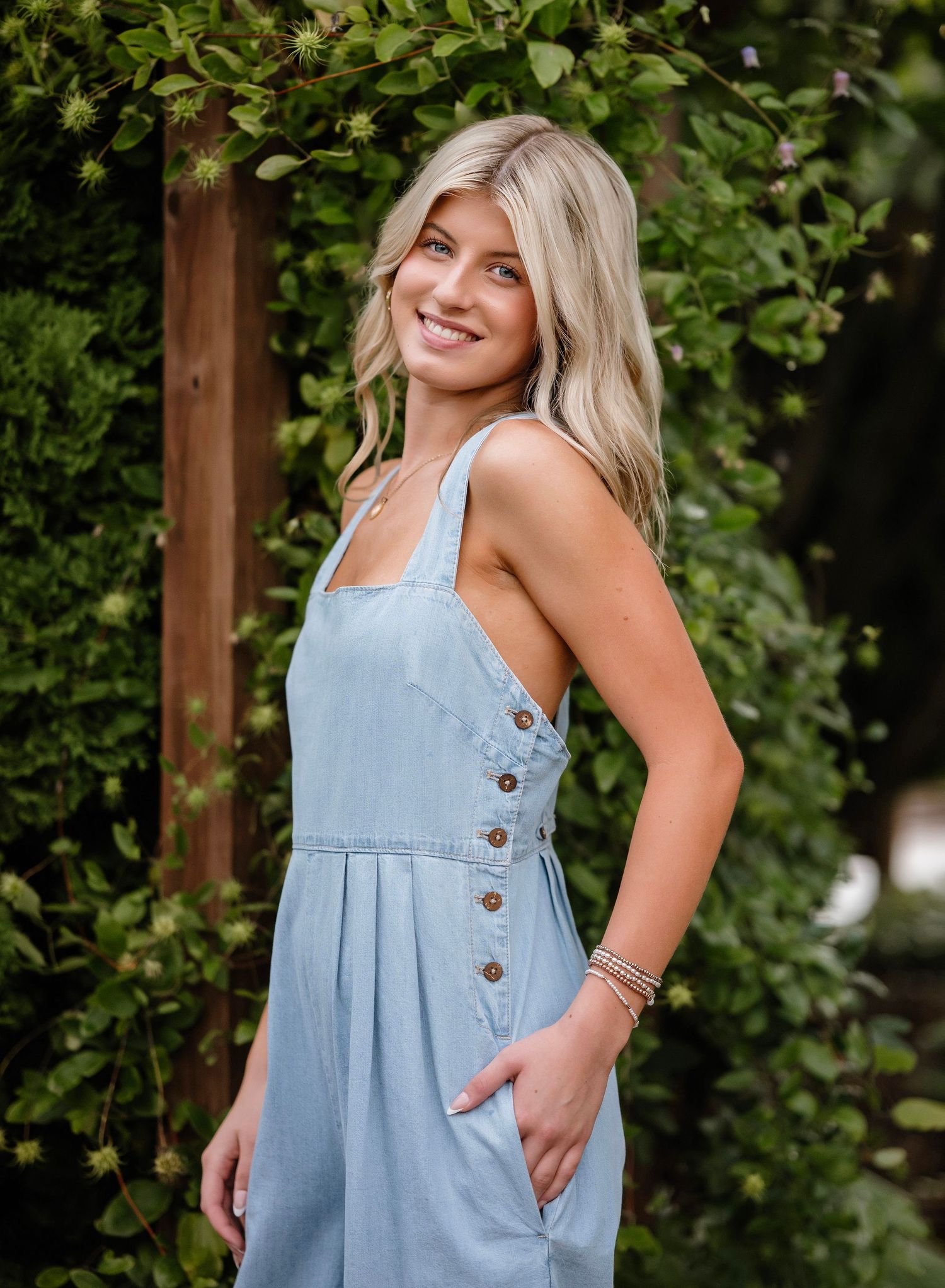 A woman wearing a light blue denim dress with button details poses outdoors against a leafy green background.