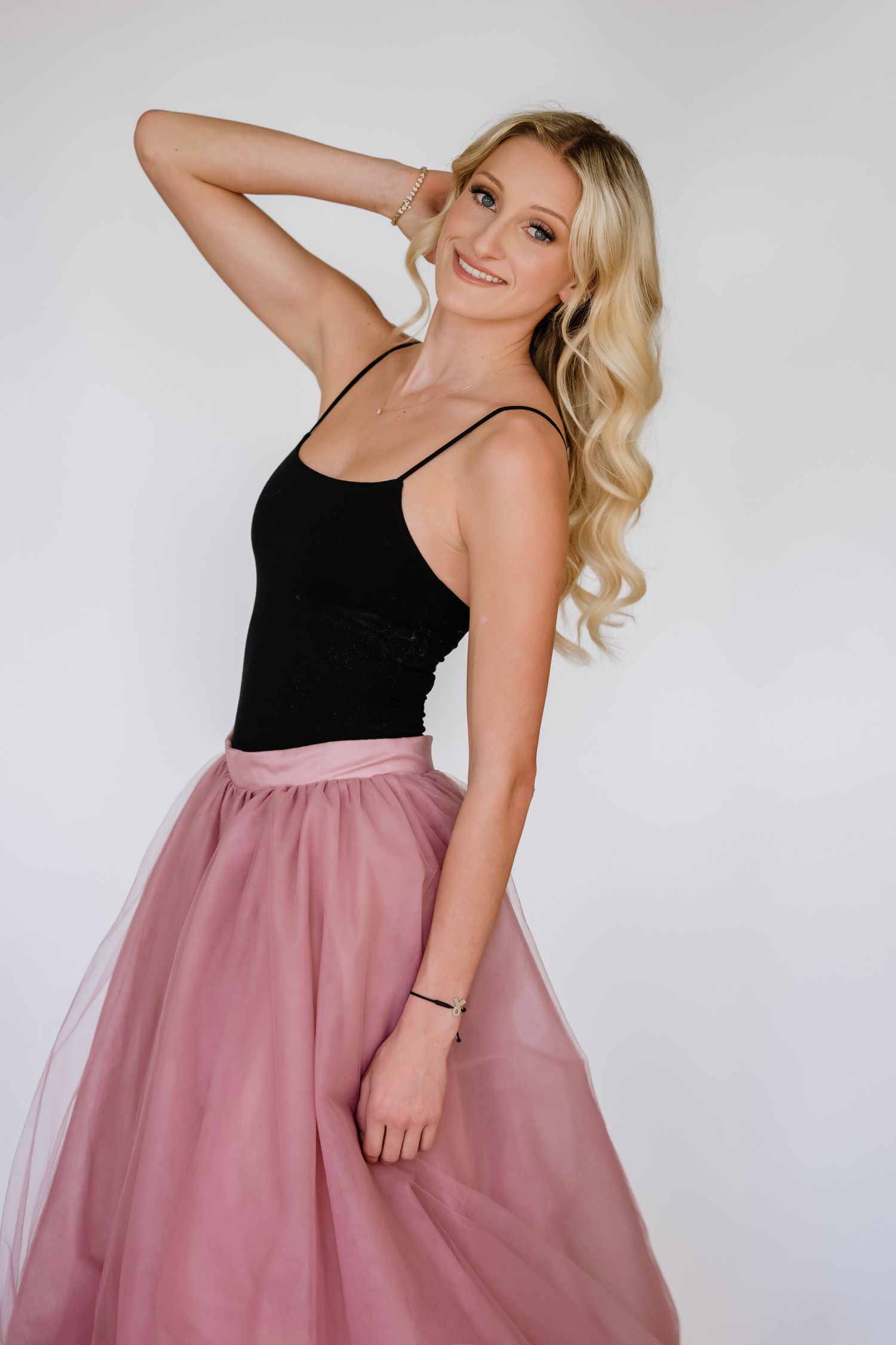 Blonde woman poses elegantly in a black top and dusty rose tulle skirt against a white studio backdrop.