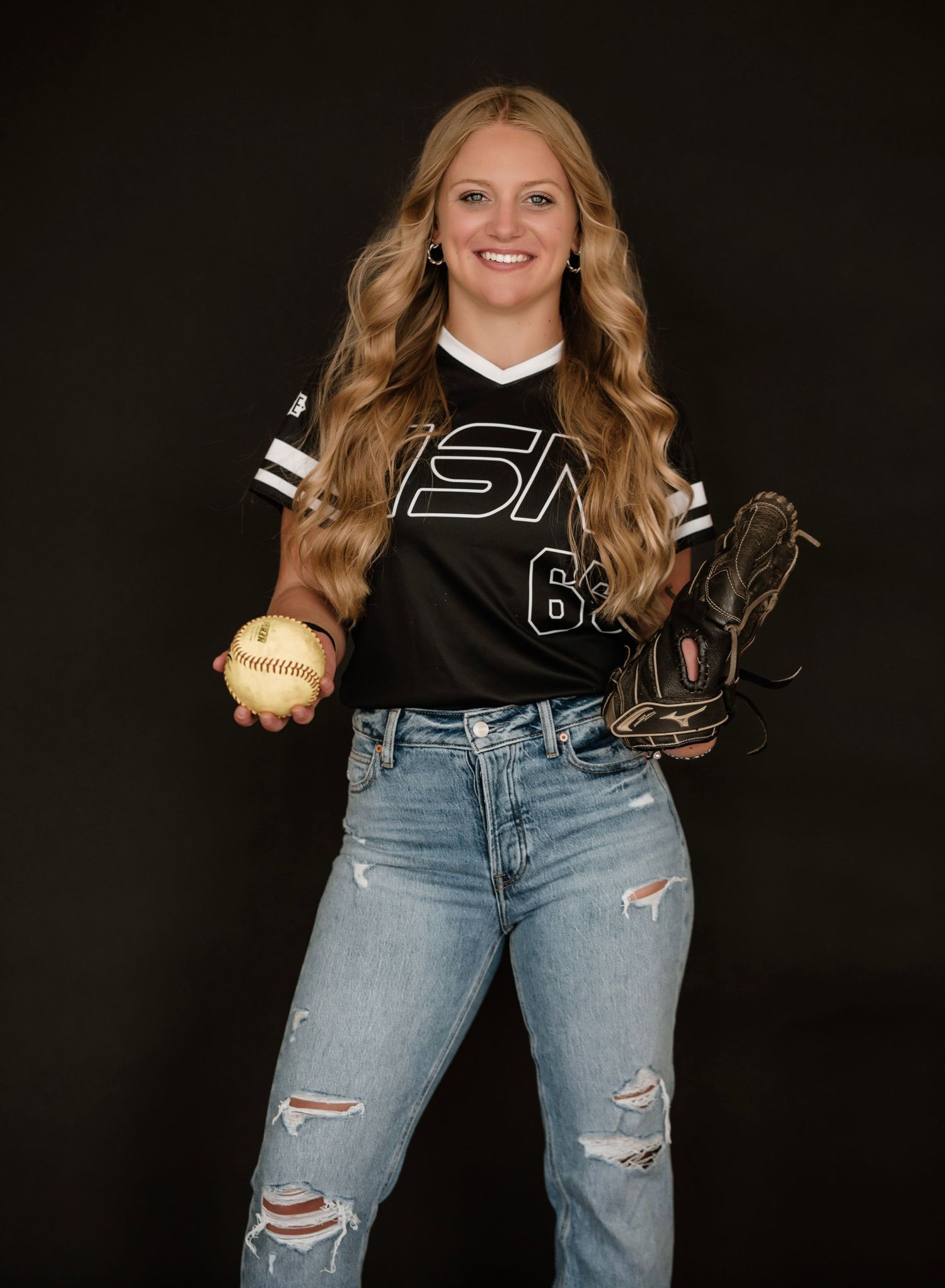 Female softball player in black jersey number 63 holding ball and glove, wearing ripped jeans, posing for portrait.
