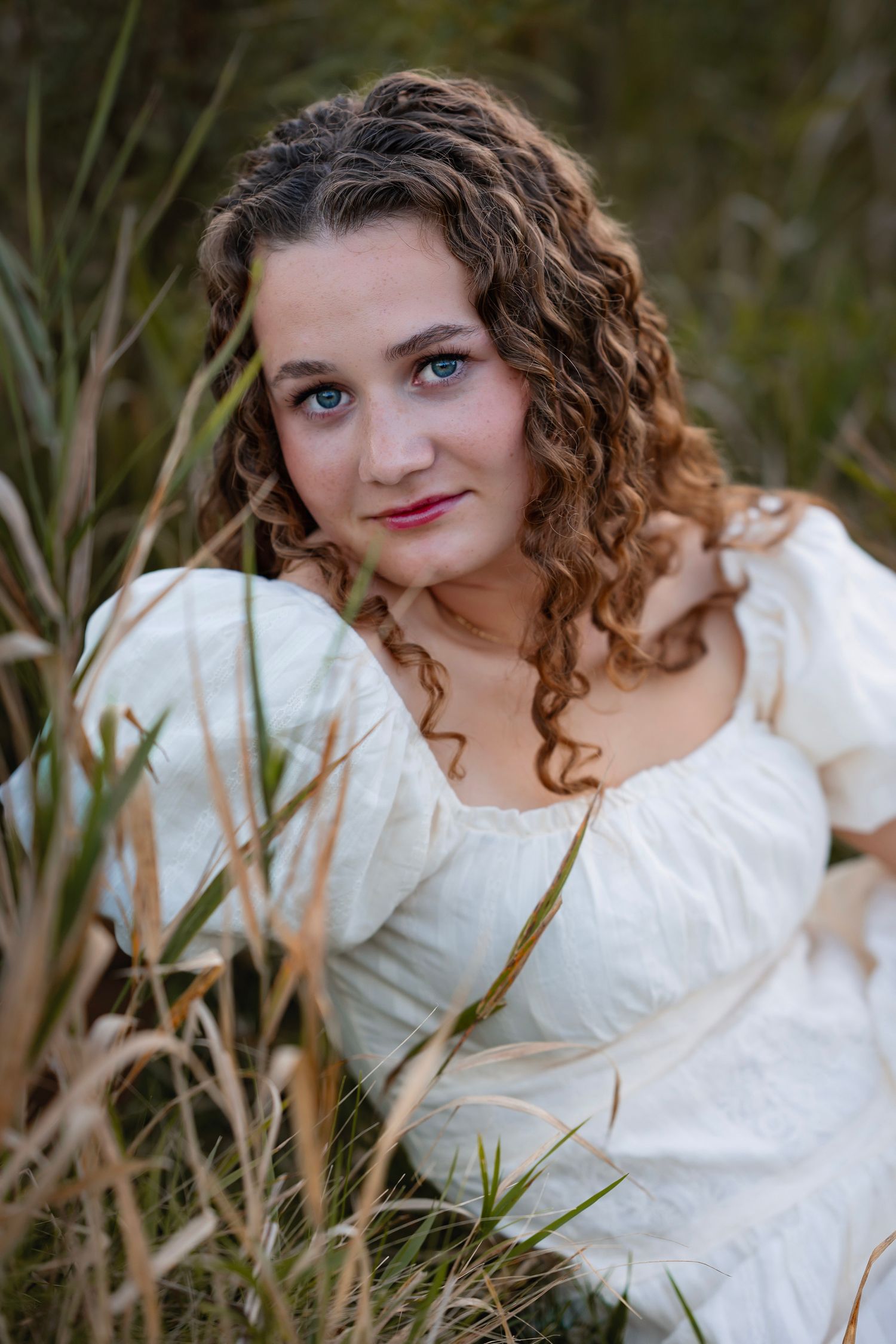 Young woman with curly hair in white dress sitting among tall grass, gazing softly at camera outdoors.