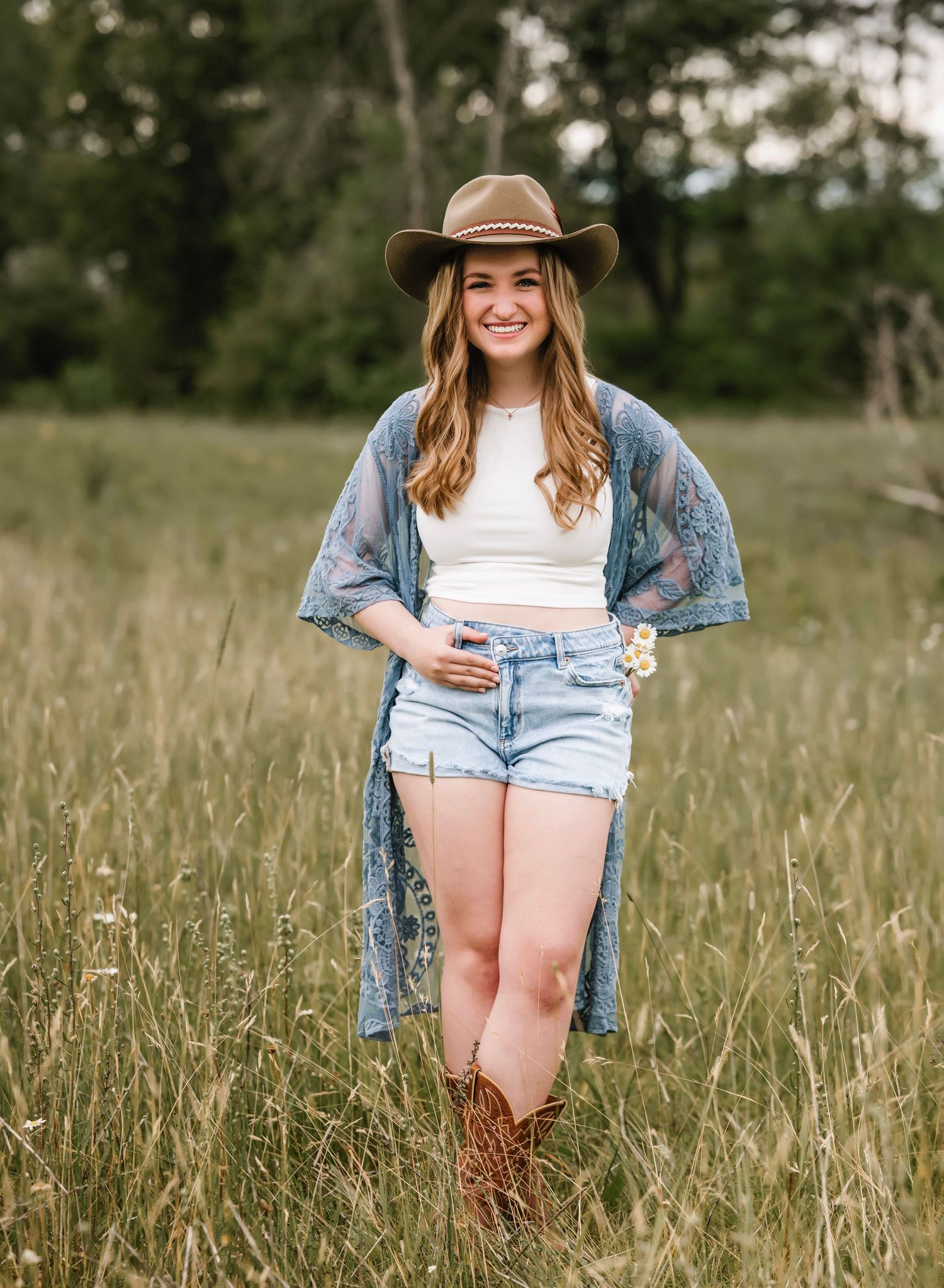 A person wearing a brown hat, denim kimono, white crop top and jean shorts stands in a tall grass field.