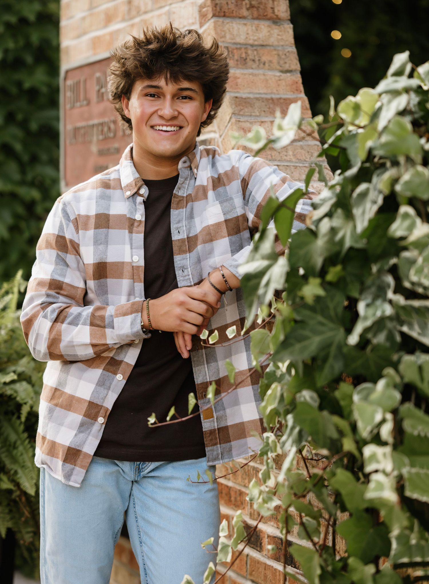 A casual outdoor portrait session featuring someone in a plaid flannel shirt and jeans leaning against a brick wall.