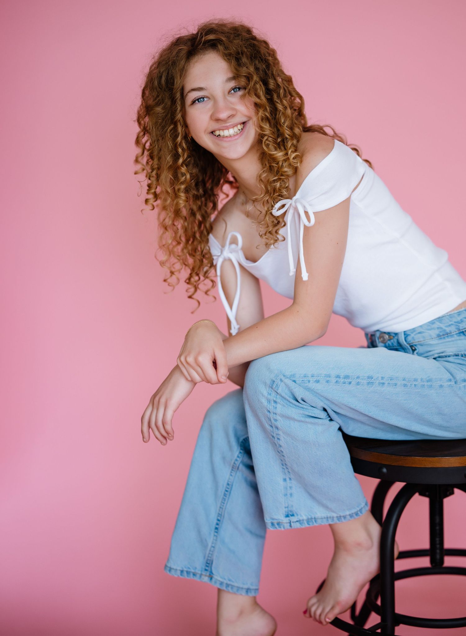 Young person in white top and light wash jeans sitting casually on stool against pink background, showing natural curly hair.