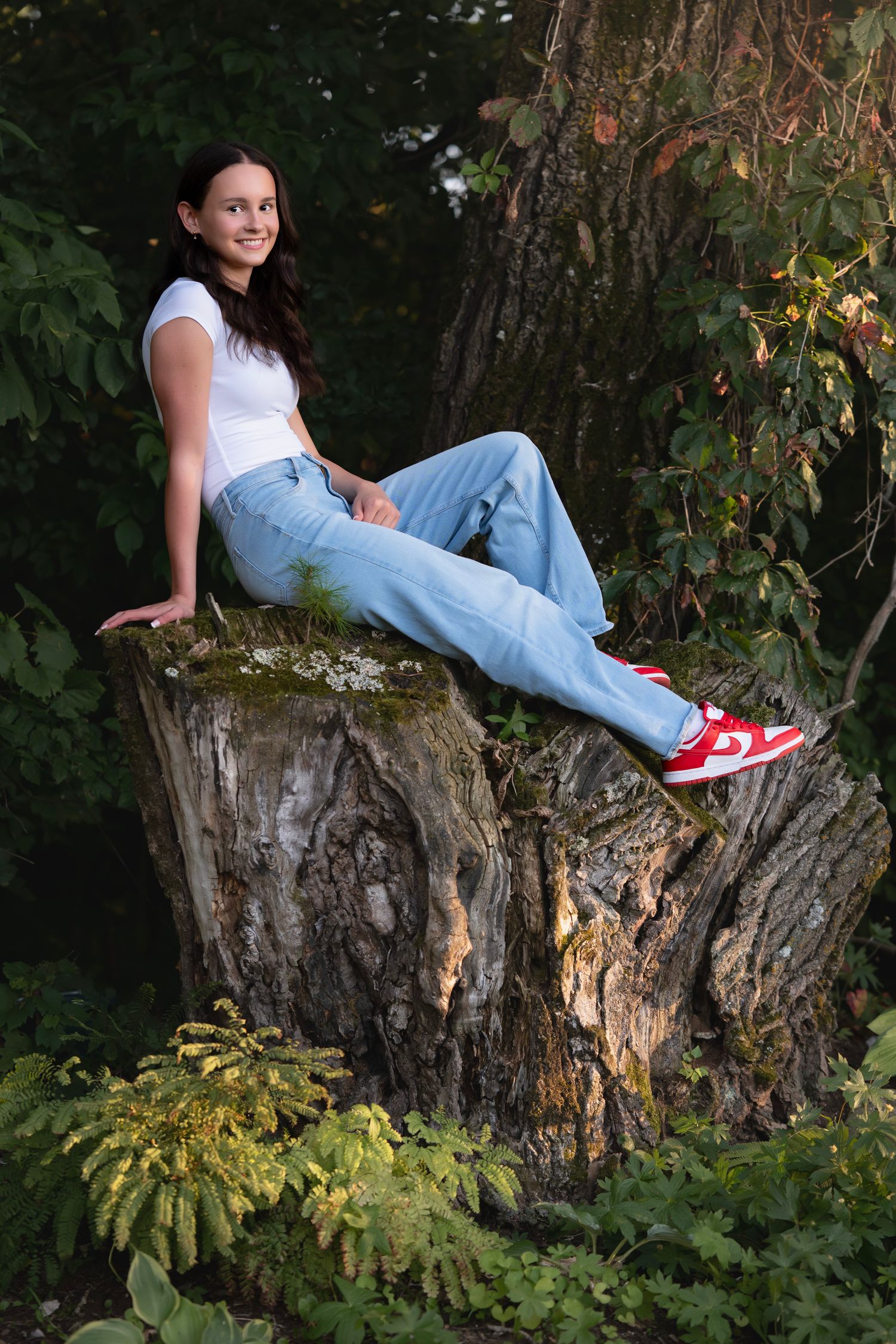 Young woman in jeans and red sneakers sitting on a large tree stump surrounded by lush green forest.