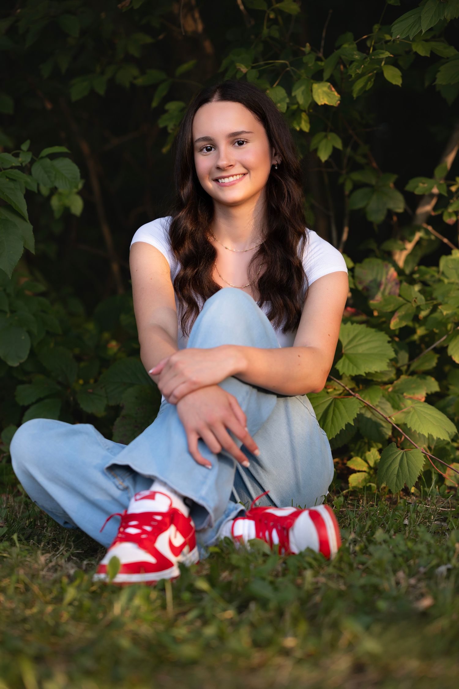 Young woman with long dark hair sits cross-legged on grass, wearing white tee, blue jeans, and red sneakers, smiling outdoors.