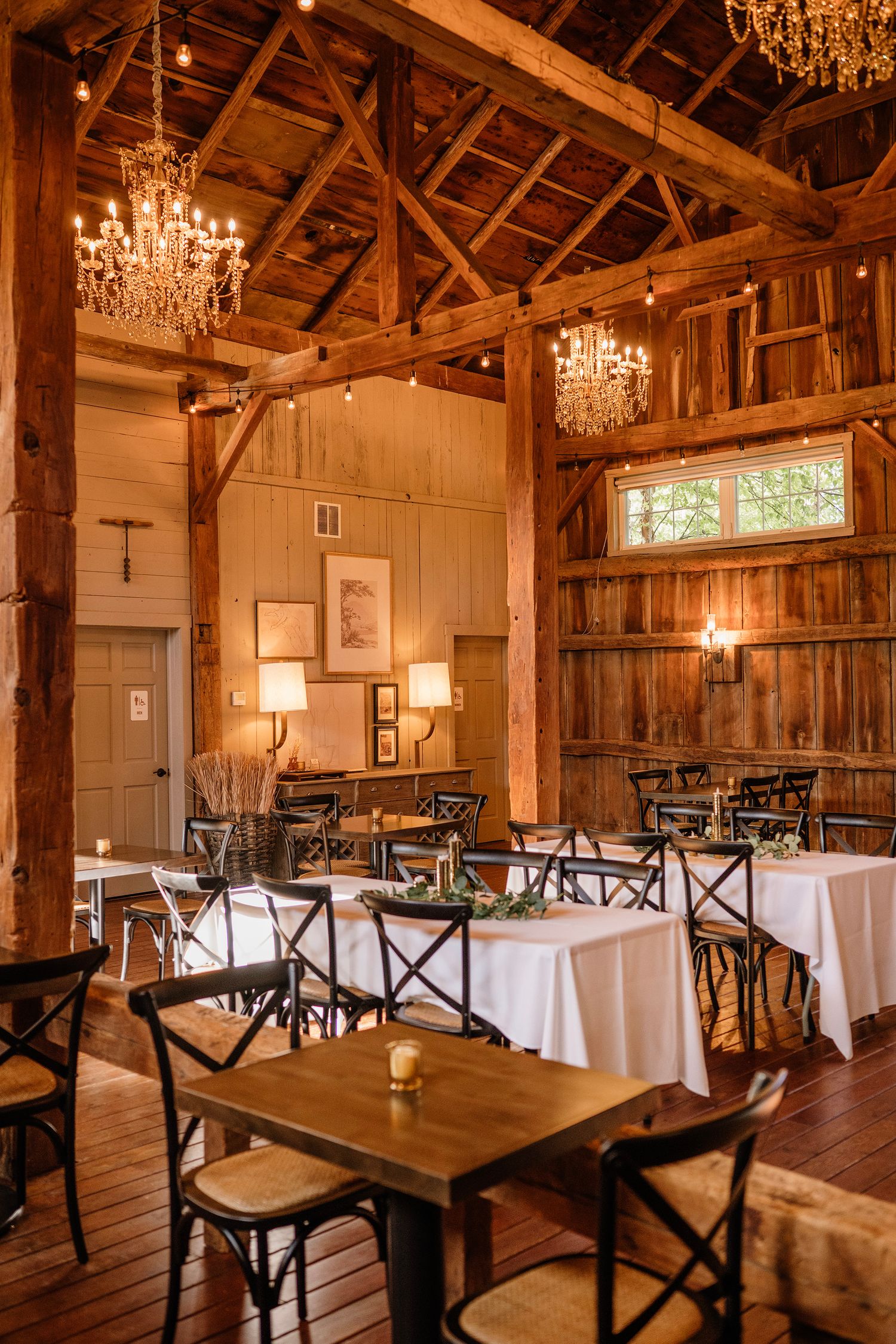 Rustic barn wedding venue with crystal chandeliers, white-clothed tables, cross-back chairs, and warm Edison lights.