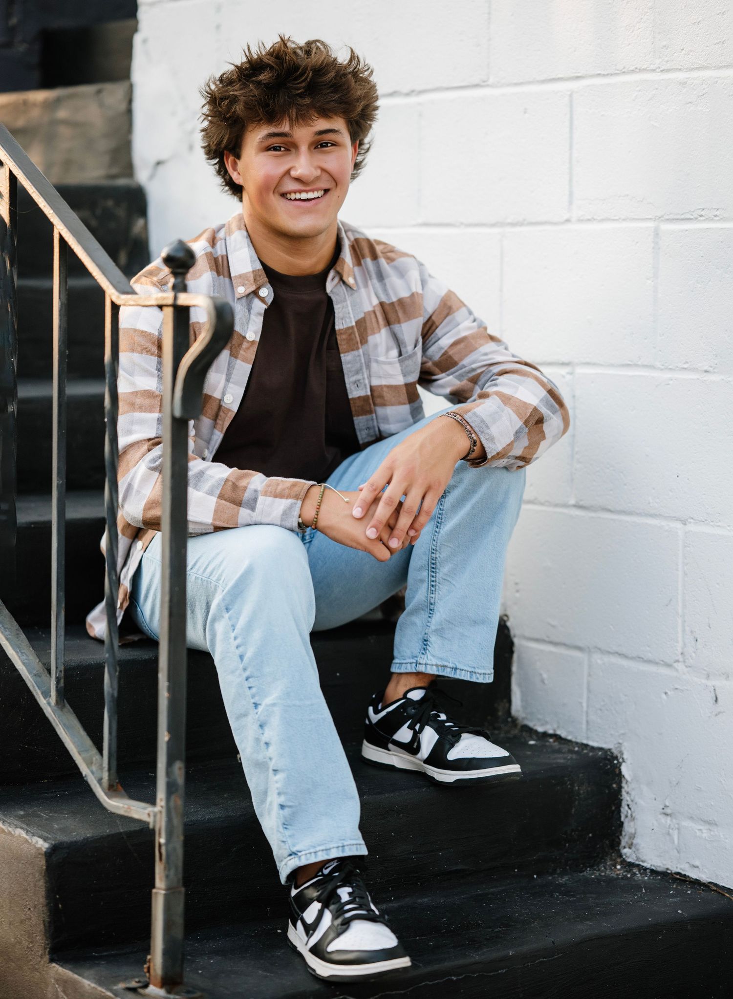 Person in a plaid shirt and jeans sits casually on black steps against a white wall in a series of candid poses.