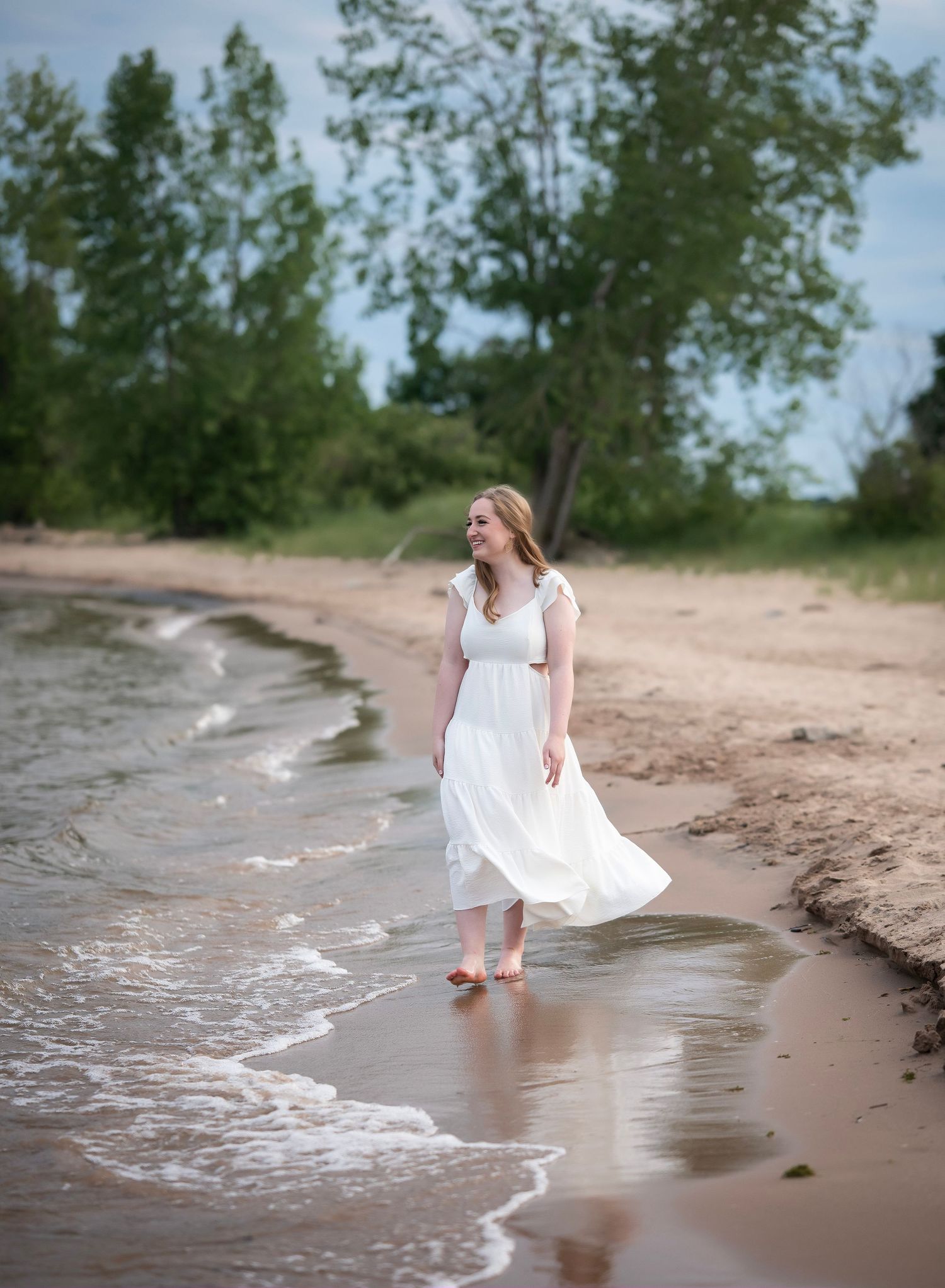 A figure in a flowing white dress walks along a sandy beach shoreline with gentle waves lapping at their feet.