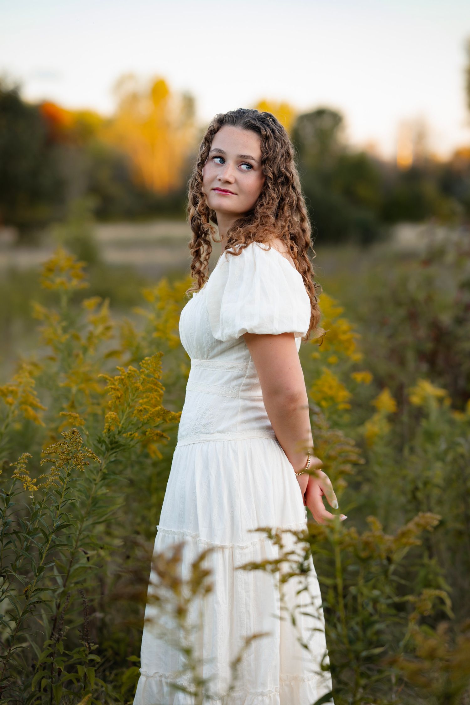 A person in a flowing white dress poses among blooming yellow goldenrod flowers during golden hour.