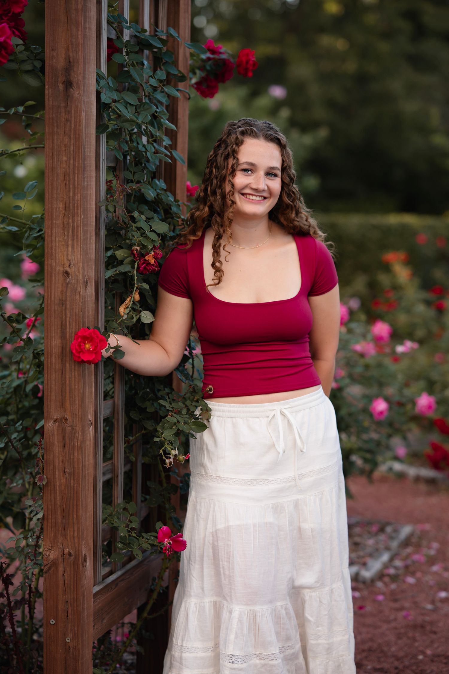 A person in a burgundy top and white tiered skirt poses in a garden setting with blooming red roses.