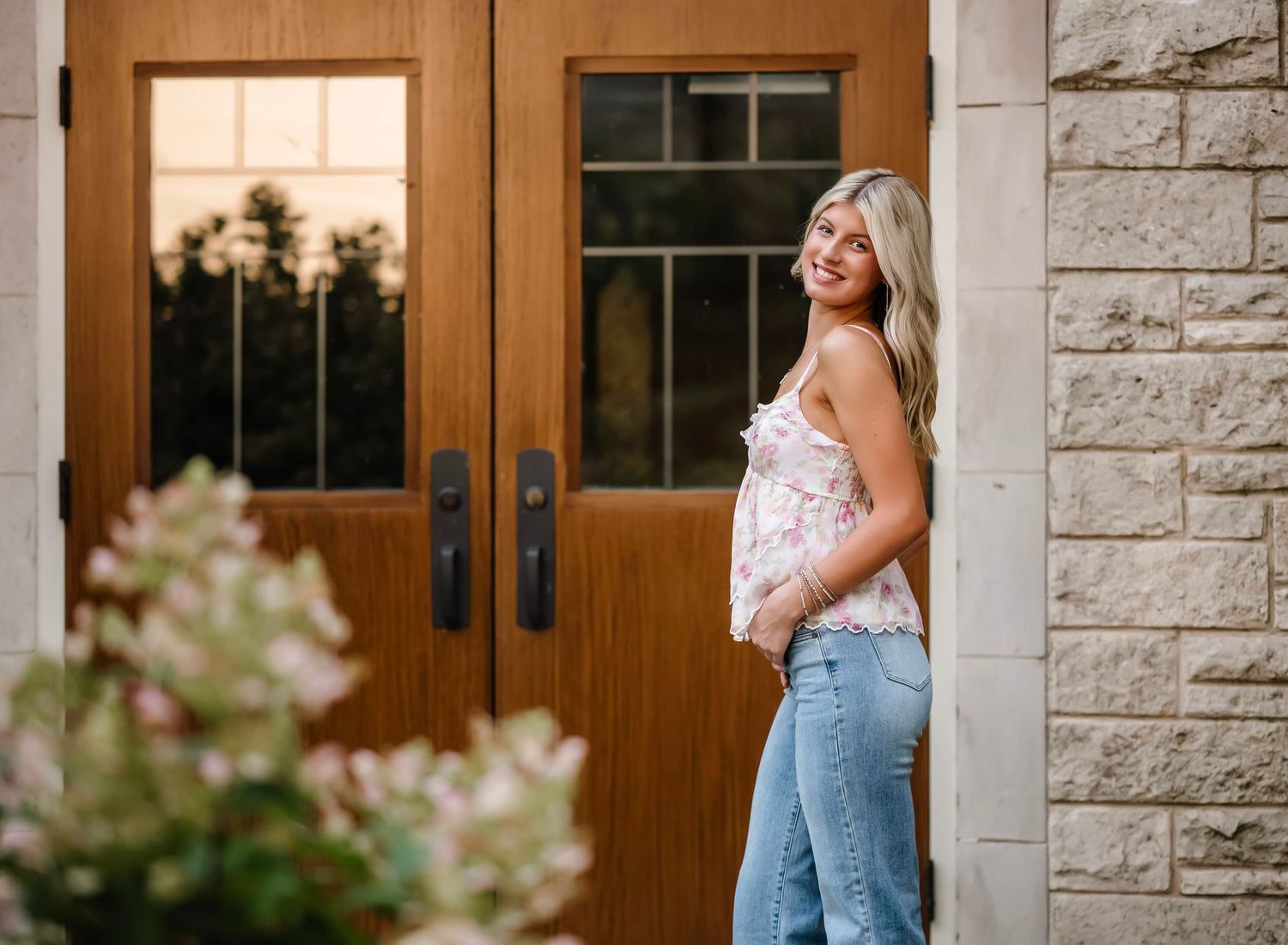A woman in overalls and white top stands in front of a rustic stone wall and wooden door.