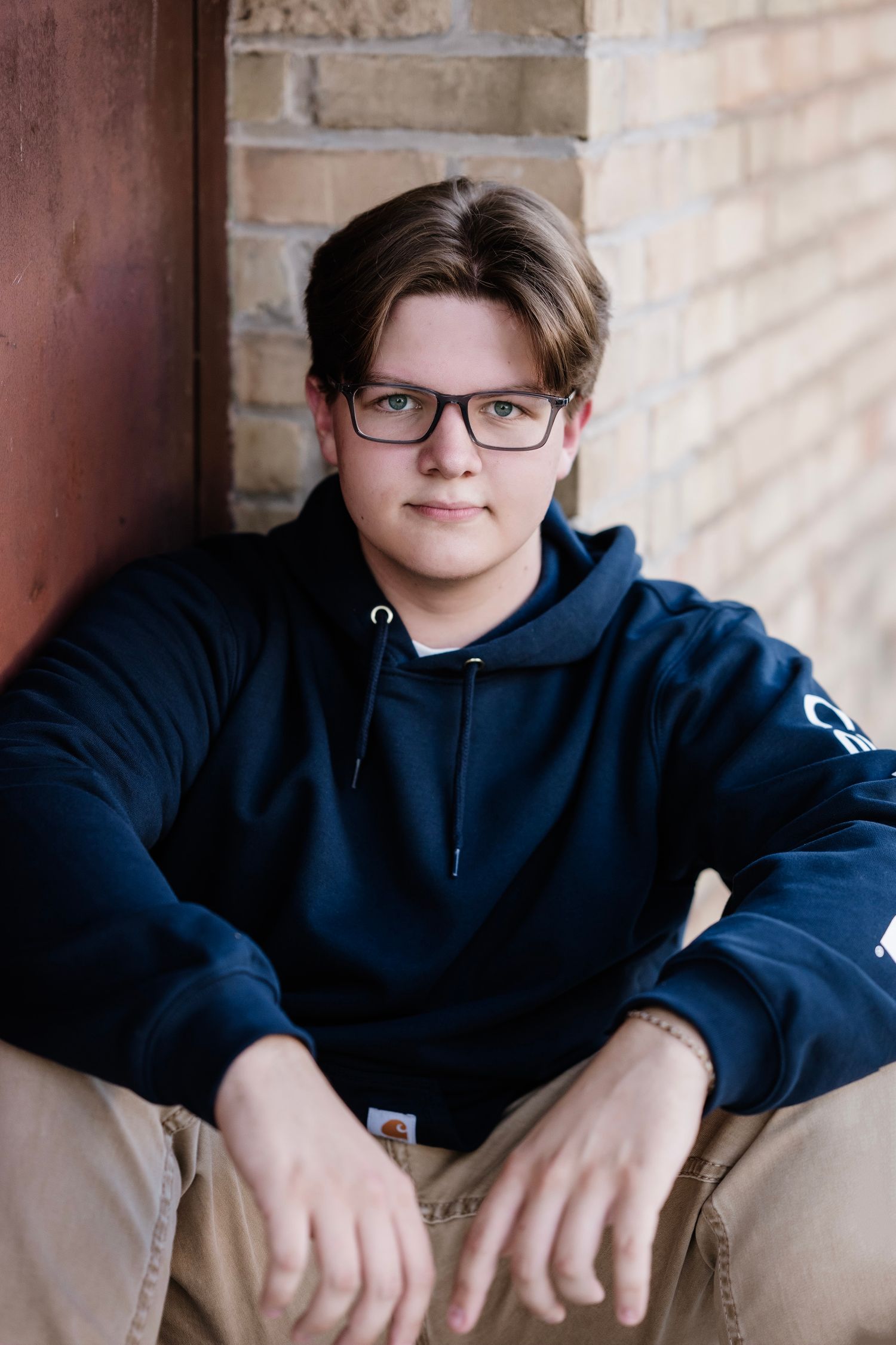 Portrait of someone wearing glasses and a navy blue hoodie sitting casually against a brick wall.