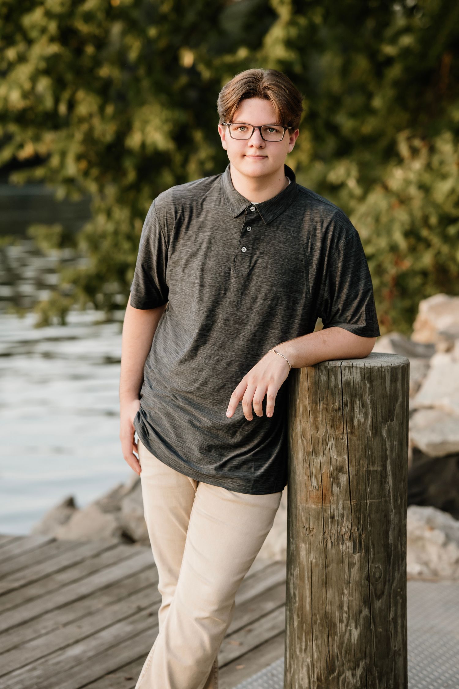 Person in a dark gray t-shirt and khaki pants poses casually against wooden posts by a waterfront.