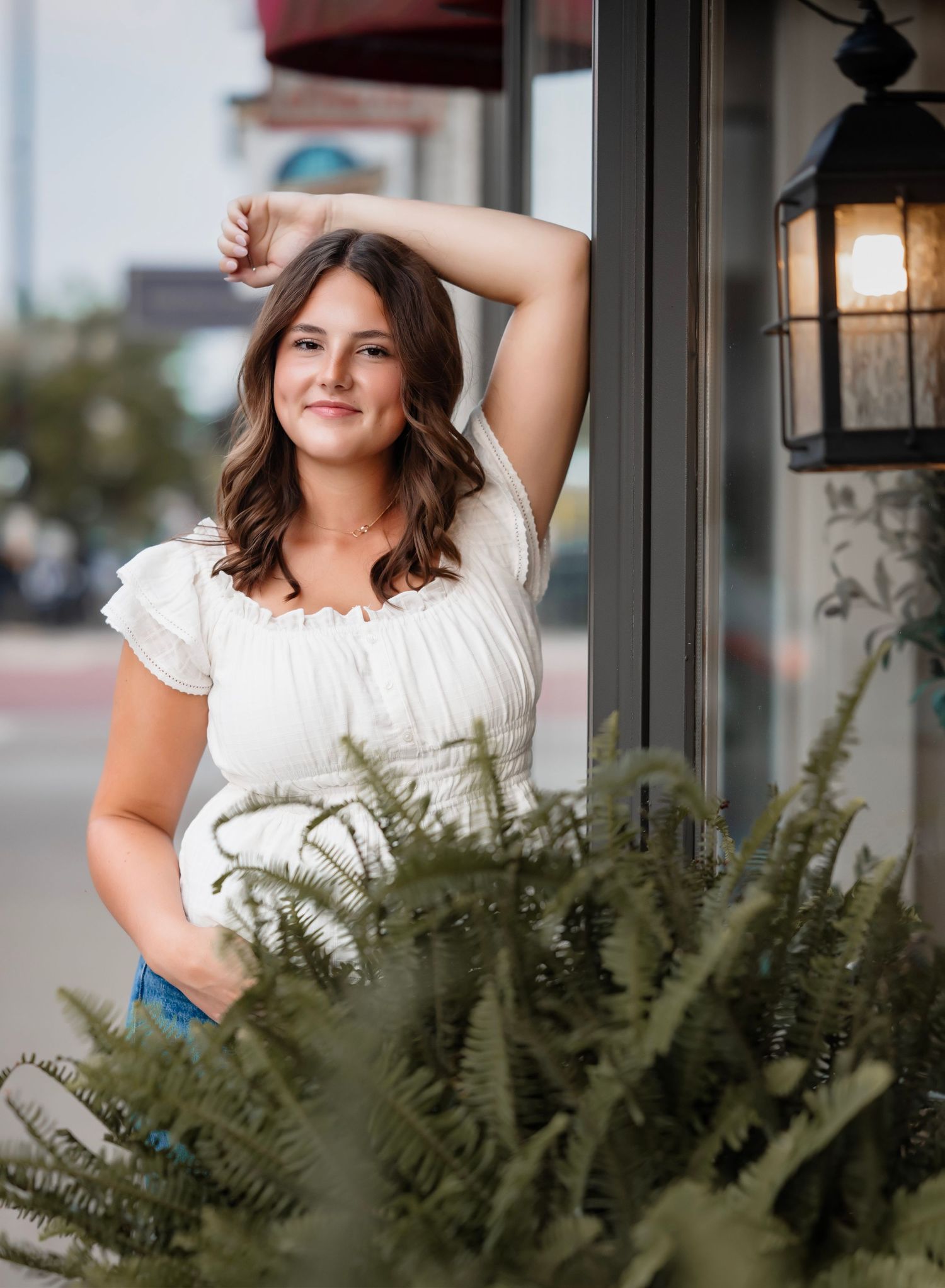 Portrait of someone in a white peasant top posing next to a decorative lantern and lush fern plant outdoors.