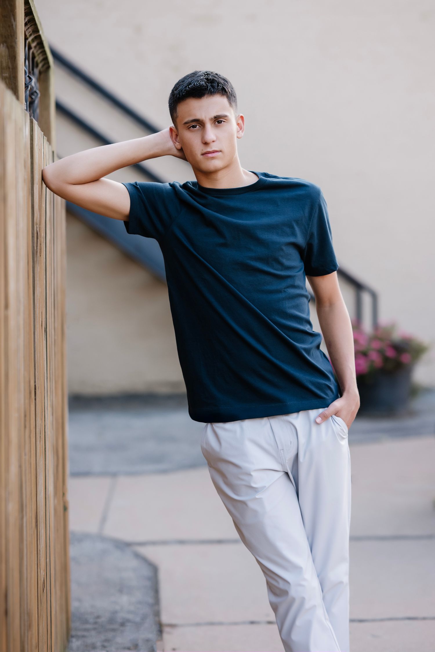 Young man in dark blue t-shirt and gray pants leaning against wooden fence outdoors.