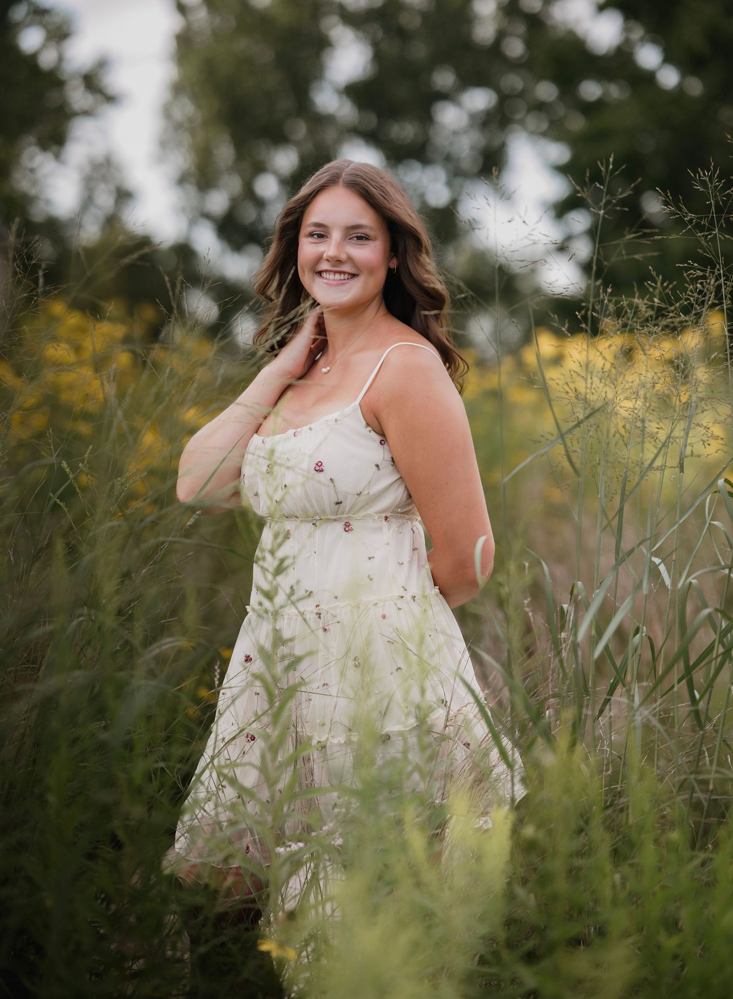 Young woman in floral sundress smiling among wildflowers in a lush green field during golden hour.