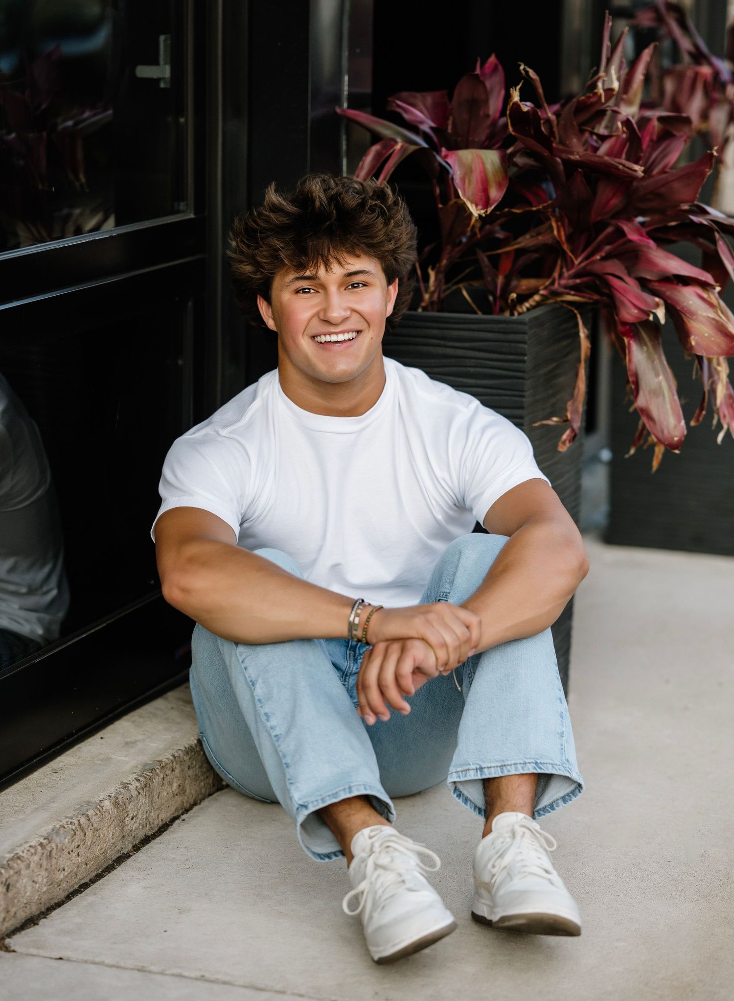 A person in a white t-shirt and light jeans sits casually on a concrete ledge with reddish plants in the background.