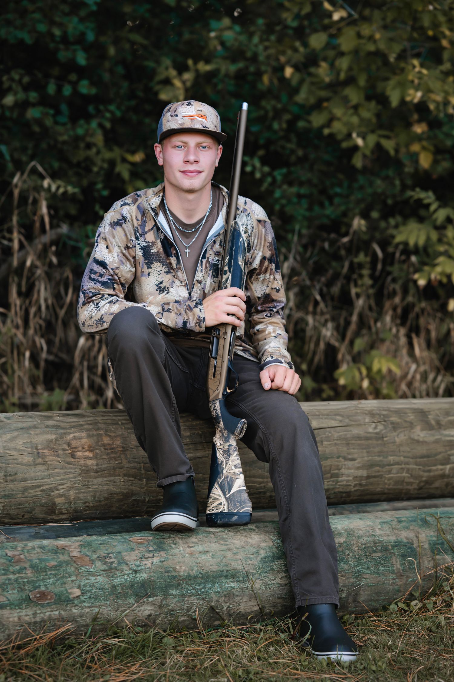 Hunter in camouflage clothing sits on fallen log holding rifle during outdoor photo session.