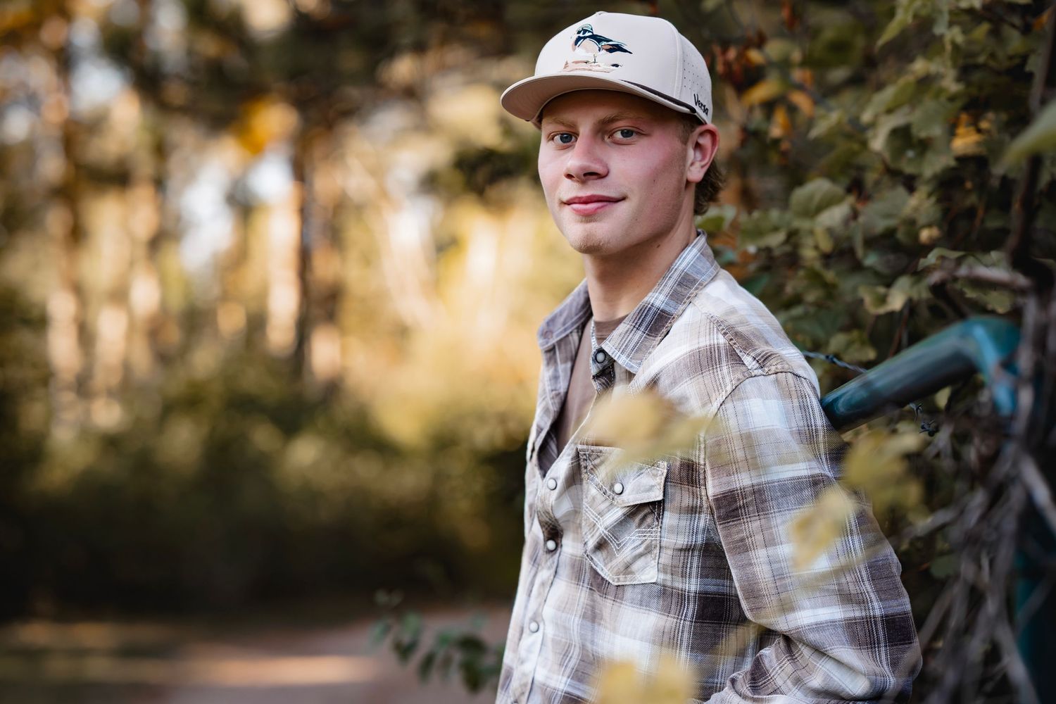 Outdoor portrait of someone wearing a plaid shirt and baseball cap in warm natural lighting with trees in background.