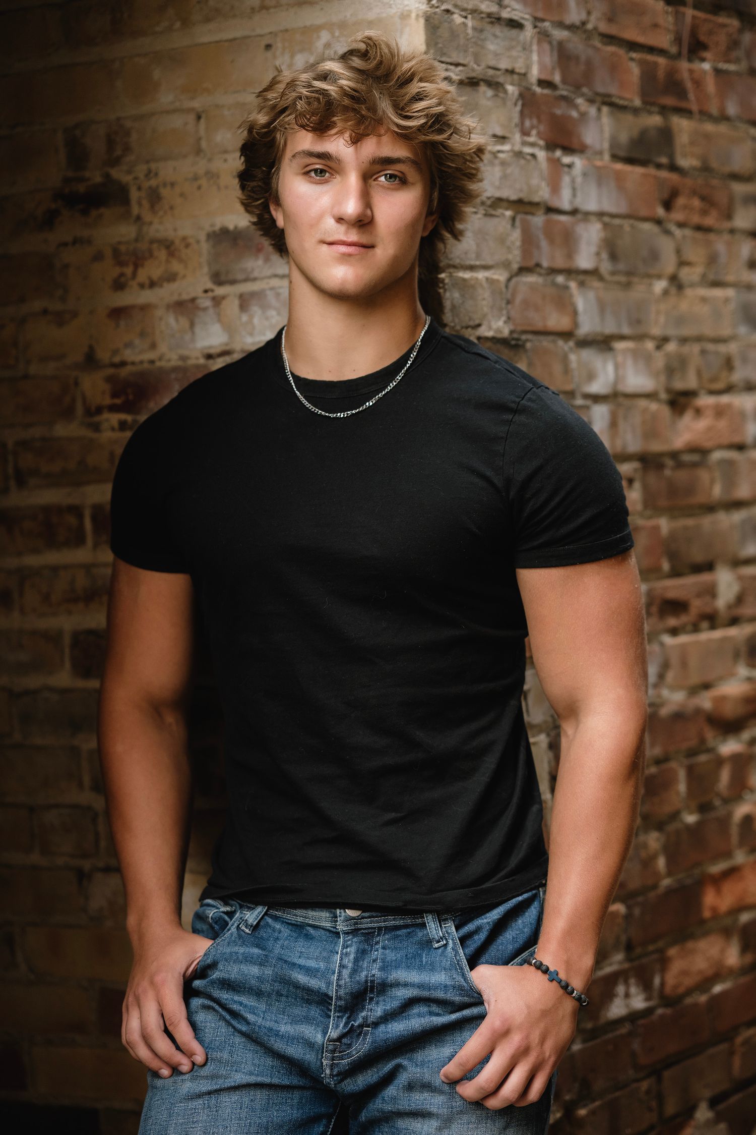 A young person in a black t-shirt poses against a rustic brick wall in a casual portrait series.