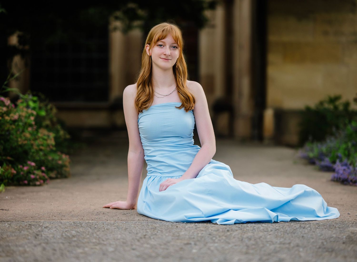 A person in a light blue formal dress sits gracefully on the ground in a garden setting with columns and flowers.