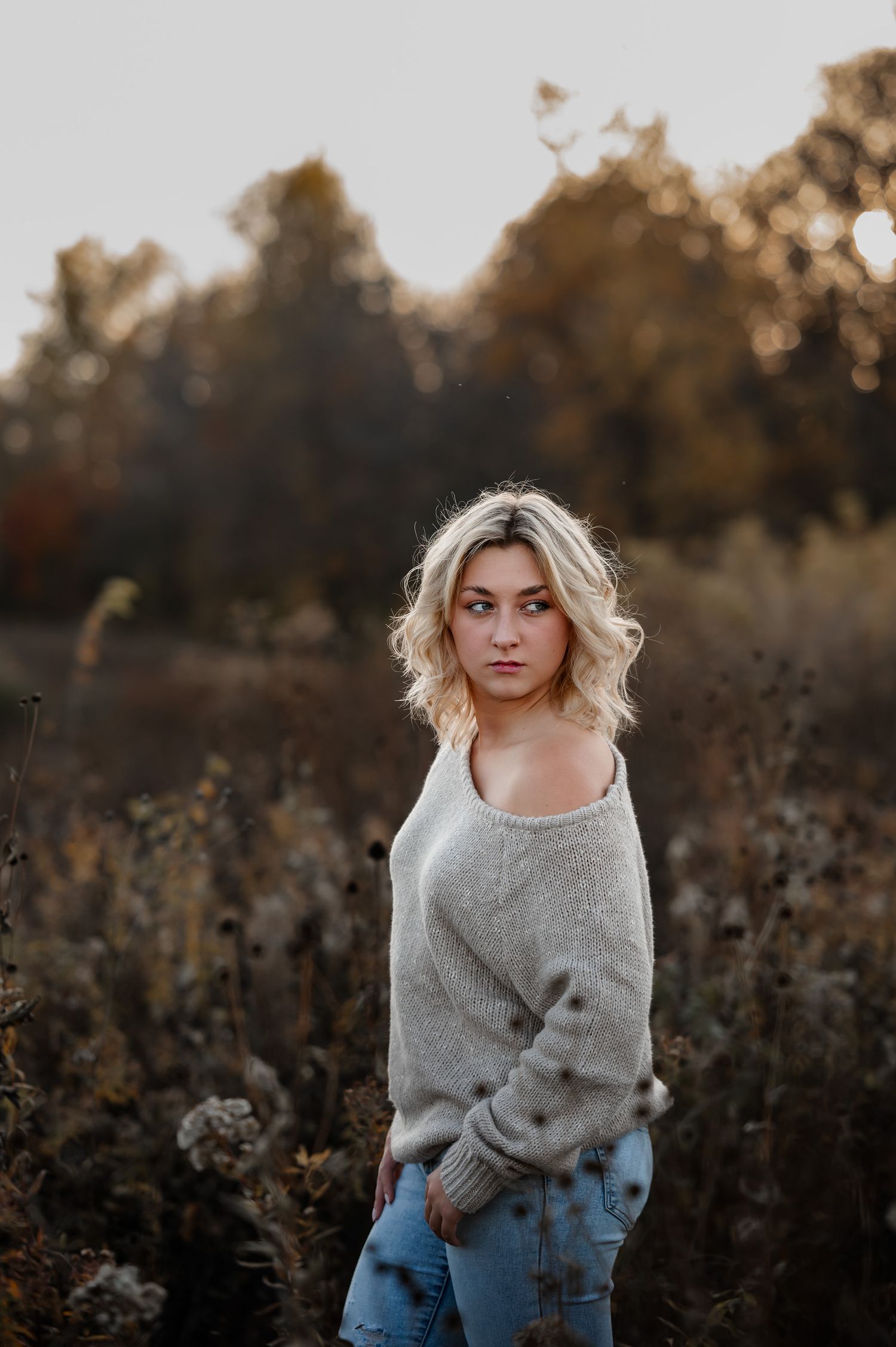 Young blonde woman in gray sweater and jeans standing in autumn field with bokeh trees background.