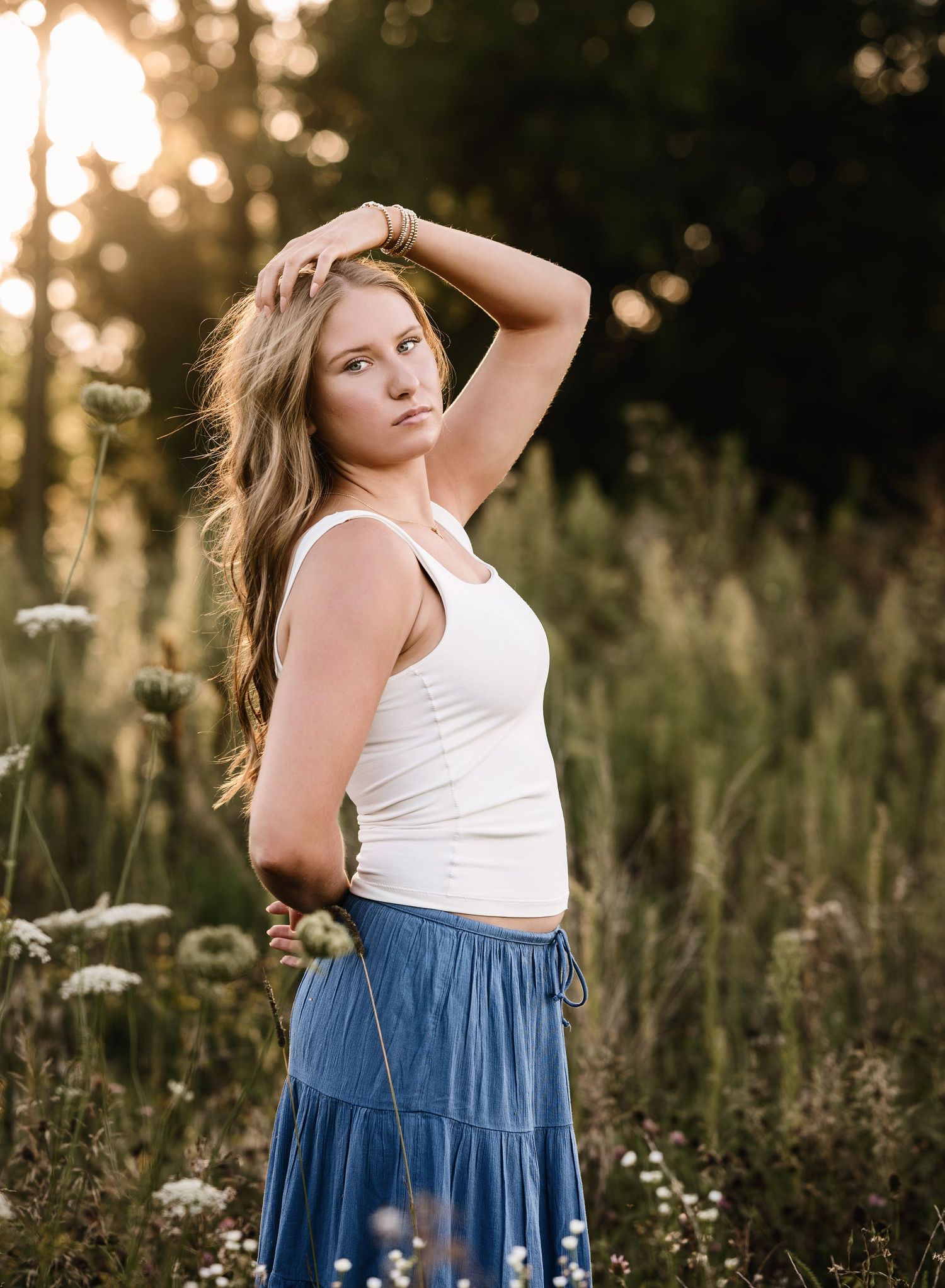 Young woman in white tank top and blue skirt poses in a sunlit wildflower meadow with golden hour light.