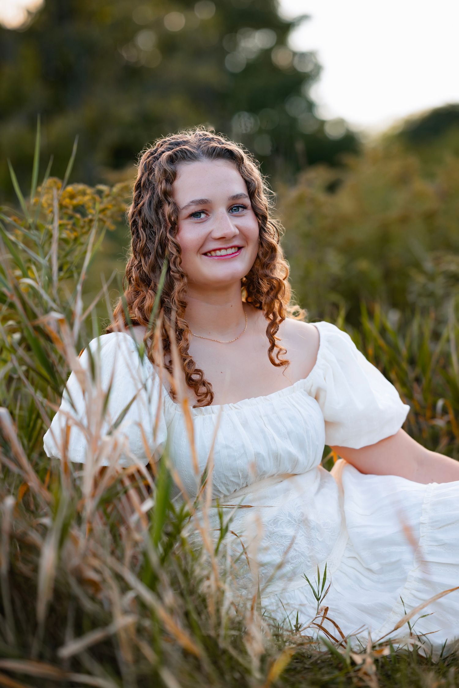 A person in a white dress poses in a field of tall dried grass during golden hour.