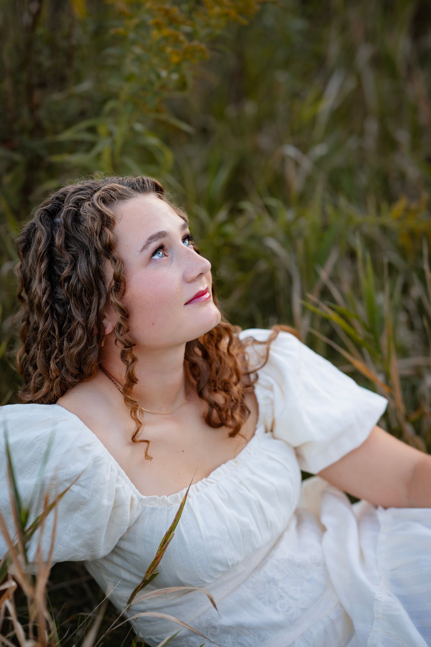 A person in a white vintage-style dress with curly hair lies in tall grass during golden hour.