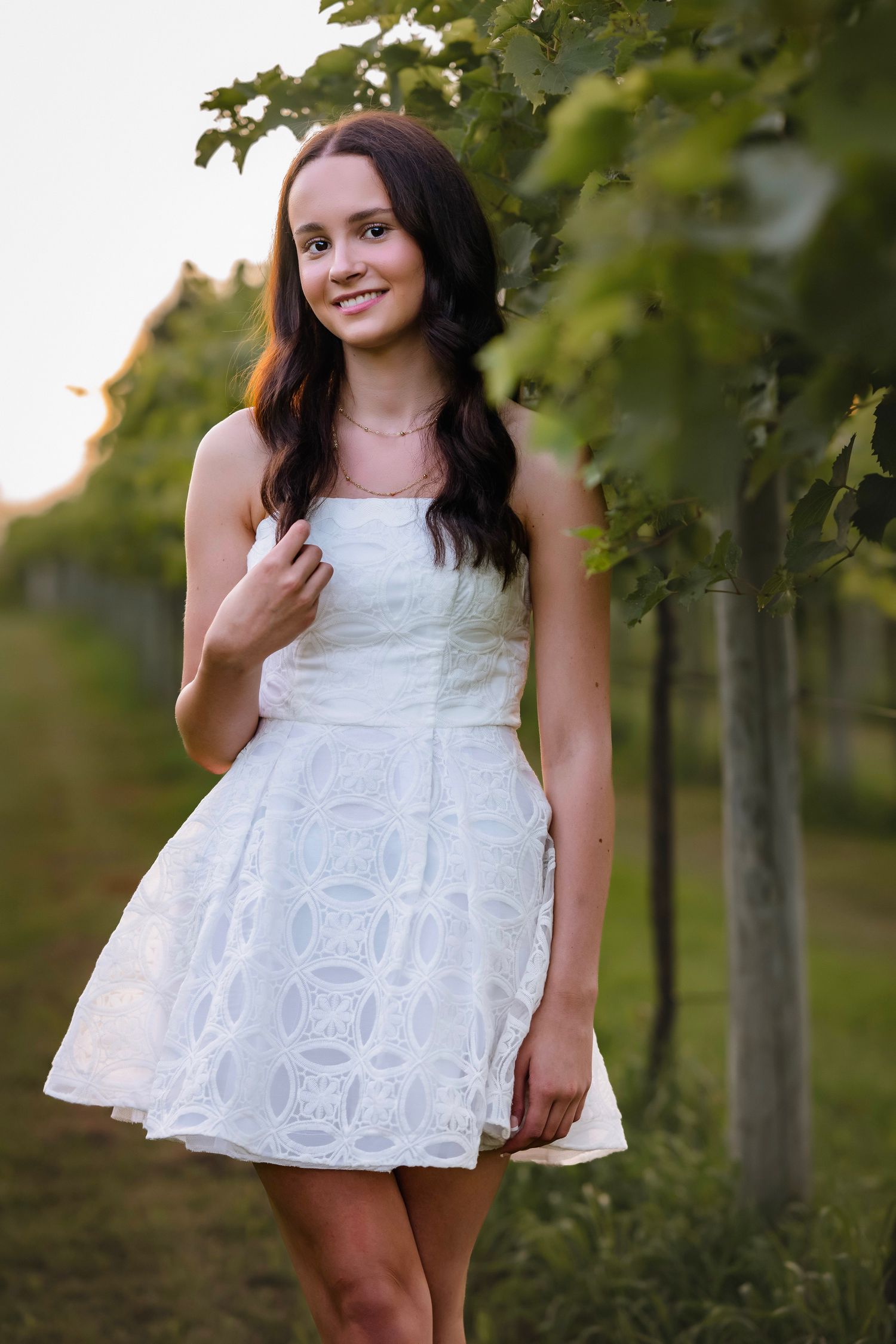 Young woman in white lace dress posing gracefully among green vineyard rows at golden hour.