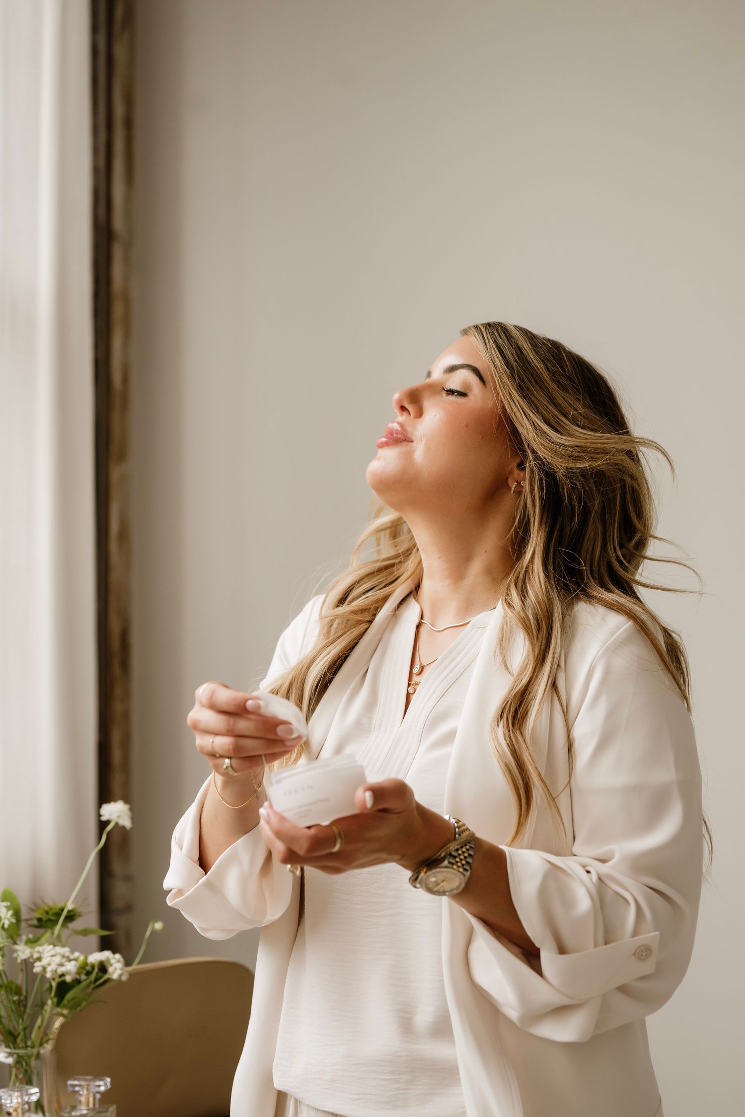 Woman in white blazer holding a cream jar with eyes closed, enjoying a skincare moment indoors.