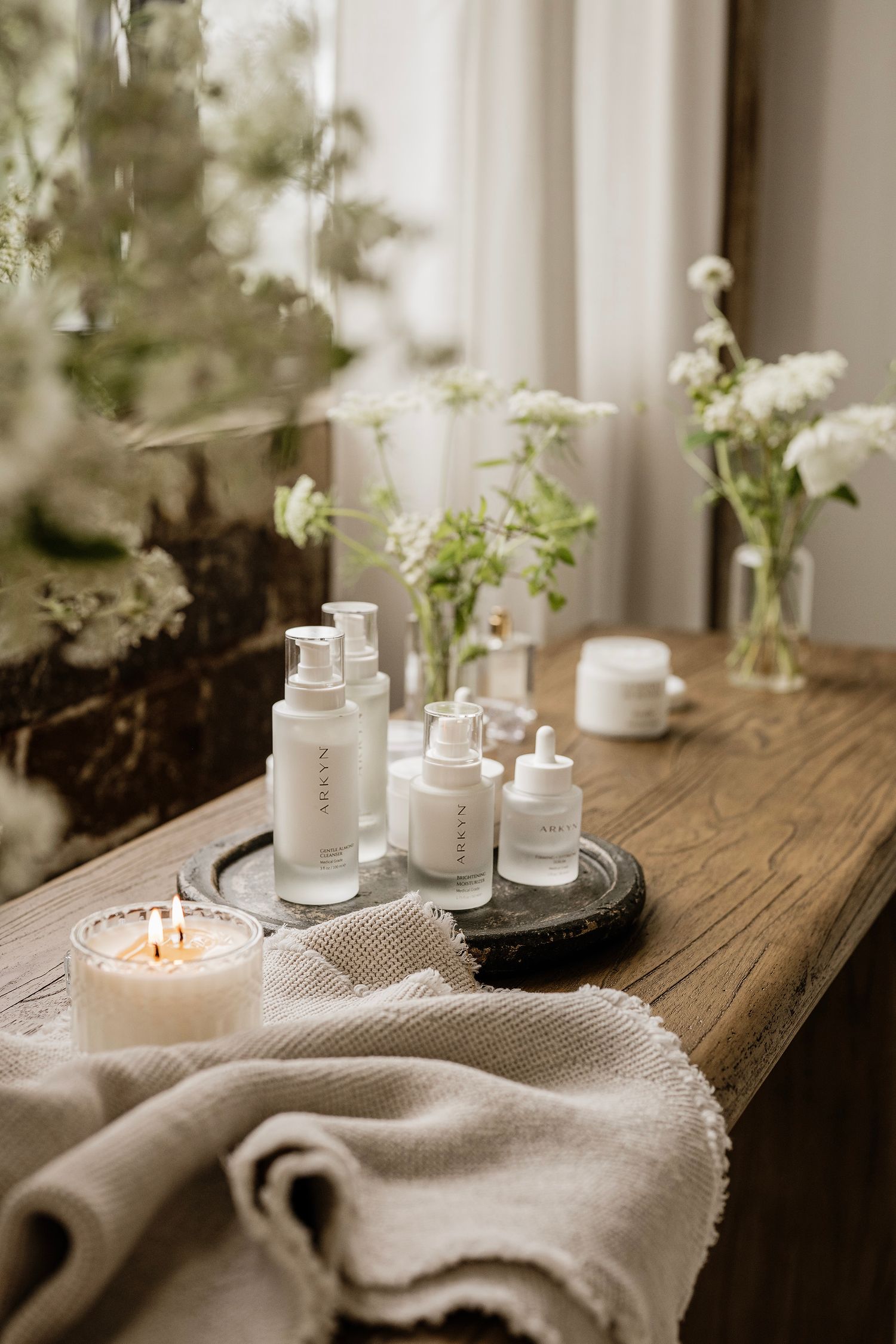 Elegant skincare products arranged on wooden shelf with white flowers, candle, and linen towel in serene bathroom setting.