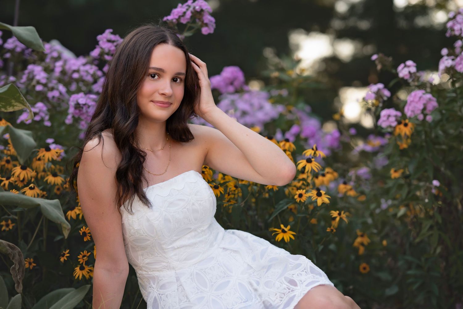 Young woman in white lace dress poses among purple and yellow wildflowers in a lush garden setting.