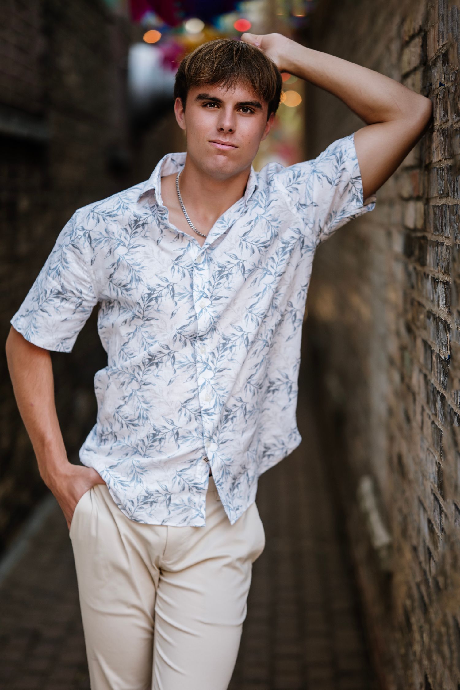 Young person in white floral shirt and khaki pants poses against stone wall in casual outdoor portrait series.